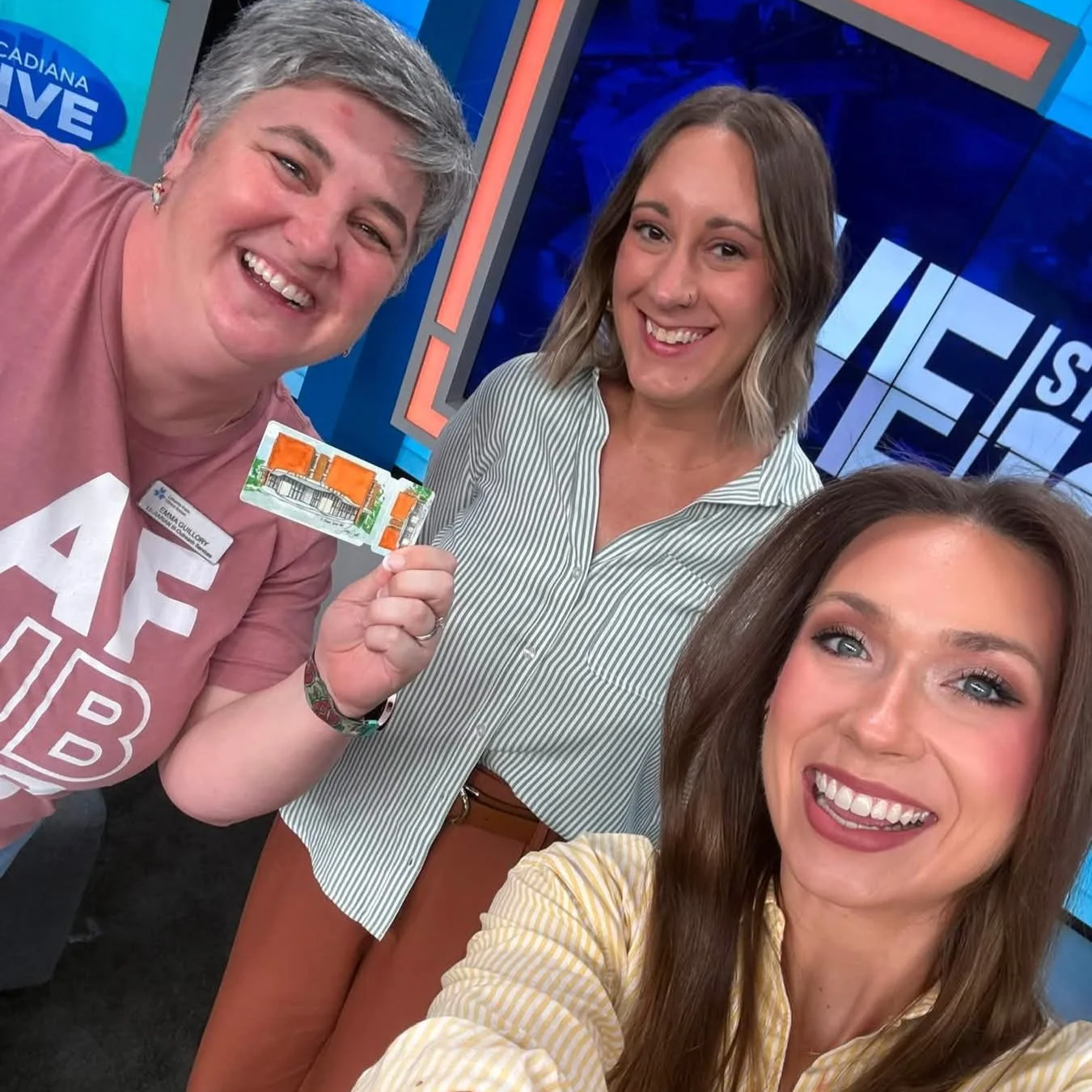 Three smiling women pose for a selfie in a television studio, with one holding a colorful illustrated card.