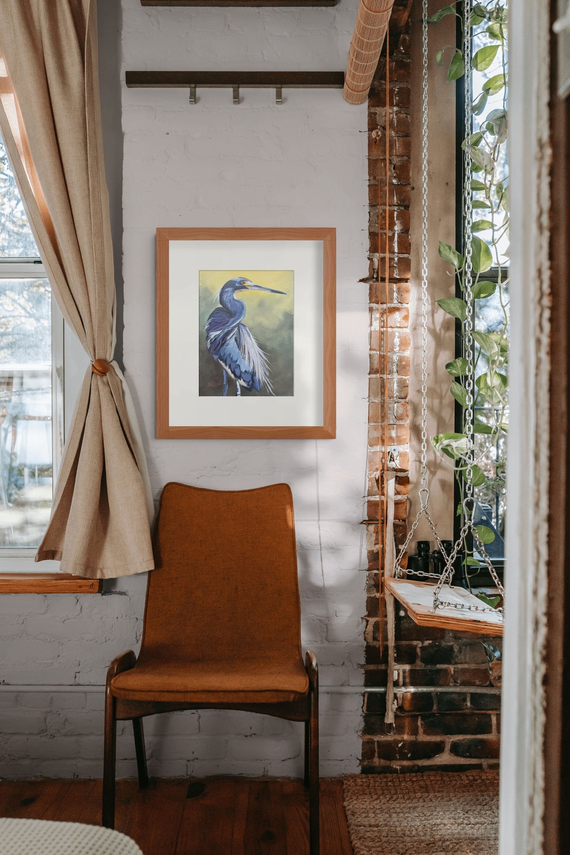 Vintage brown armchair beneath framed heron artwork in a cozy room with white brick walls, windows, curtains, and hanging plants
