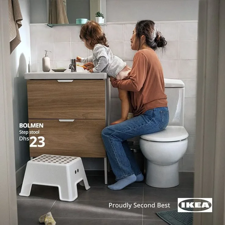 A woman sitting on the toilet holding her young child on her lap in a bathroom. The child is leaning over the bathroom sink, possibly playing or washing her hands. The scene appears to be casual and domestic.