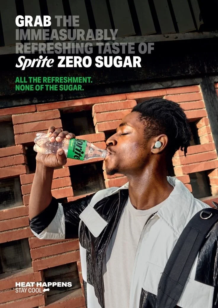 A young man with short dreadlocks drinking Sprite Zero Sugar from a plastic bottle outdoors against a brick wall. He is wearing a white and black jacket, wireless earbuds, and carrying a backpack.