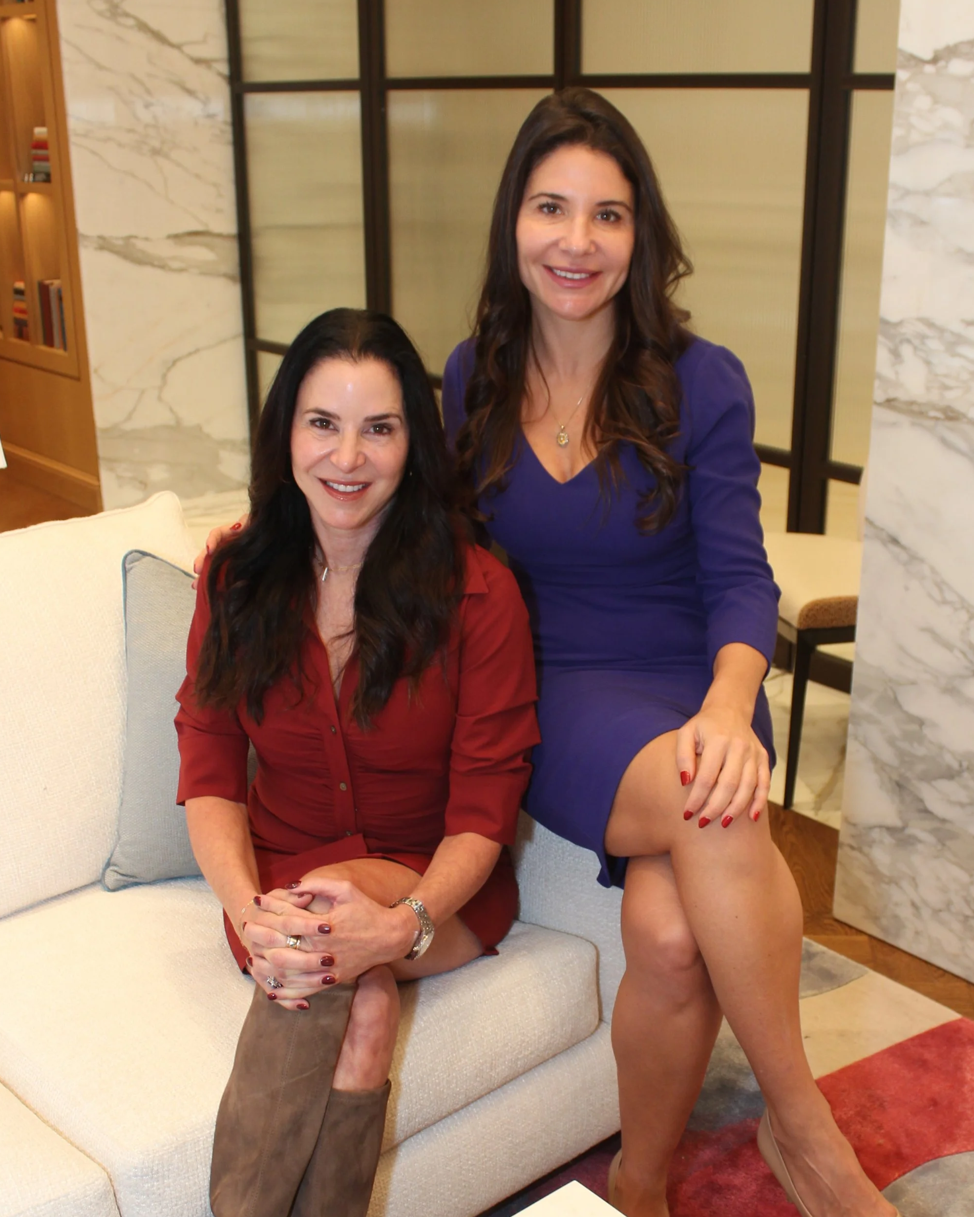 Two women smiling together in a modern living room, one seated on a sofa in a red dress and the other standing beside her in a purple dress