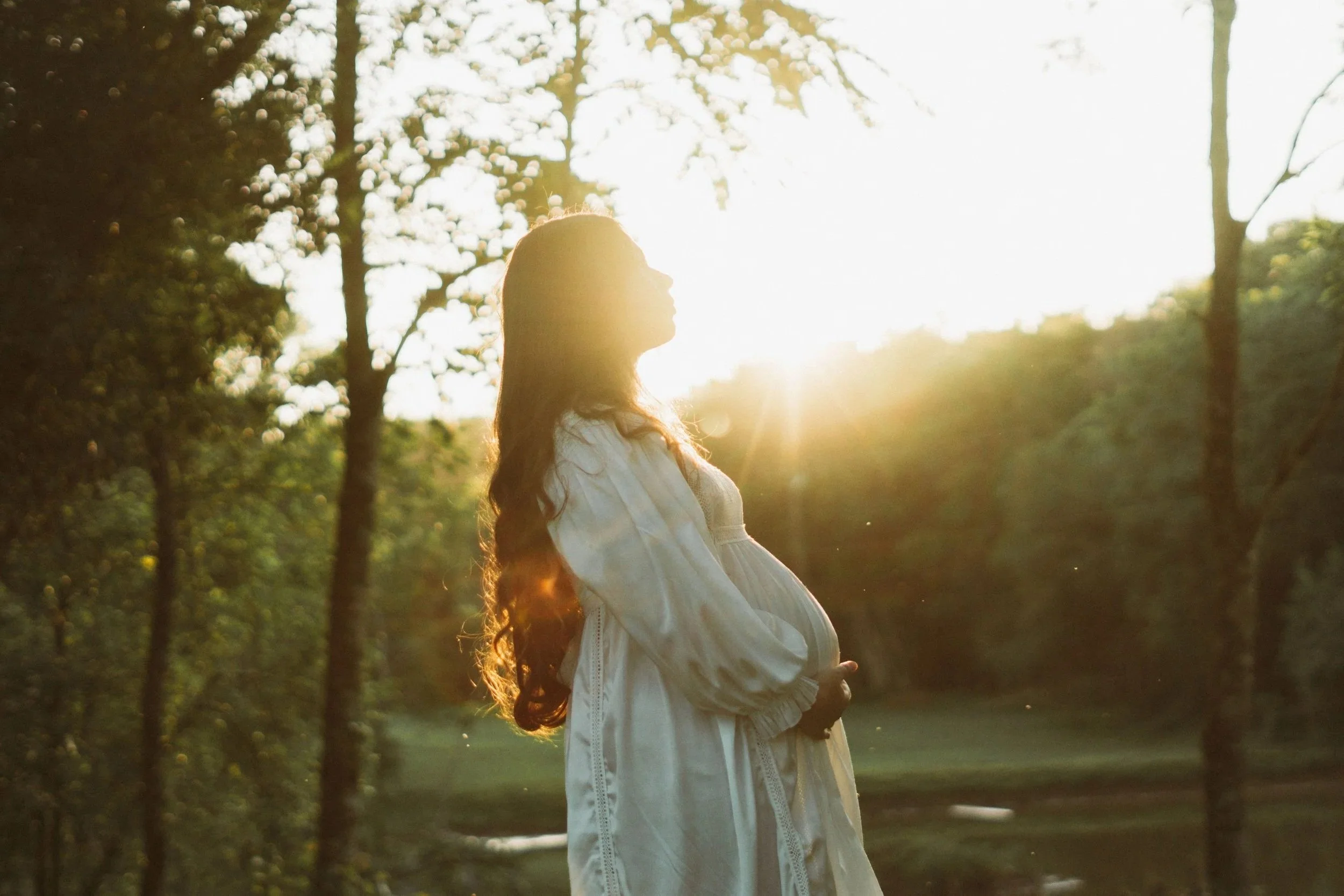 Pregnant woman in a white dress standing in a sunlit forest at golden hour