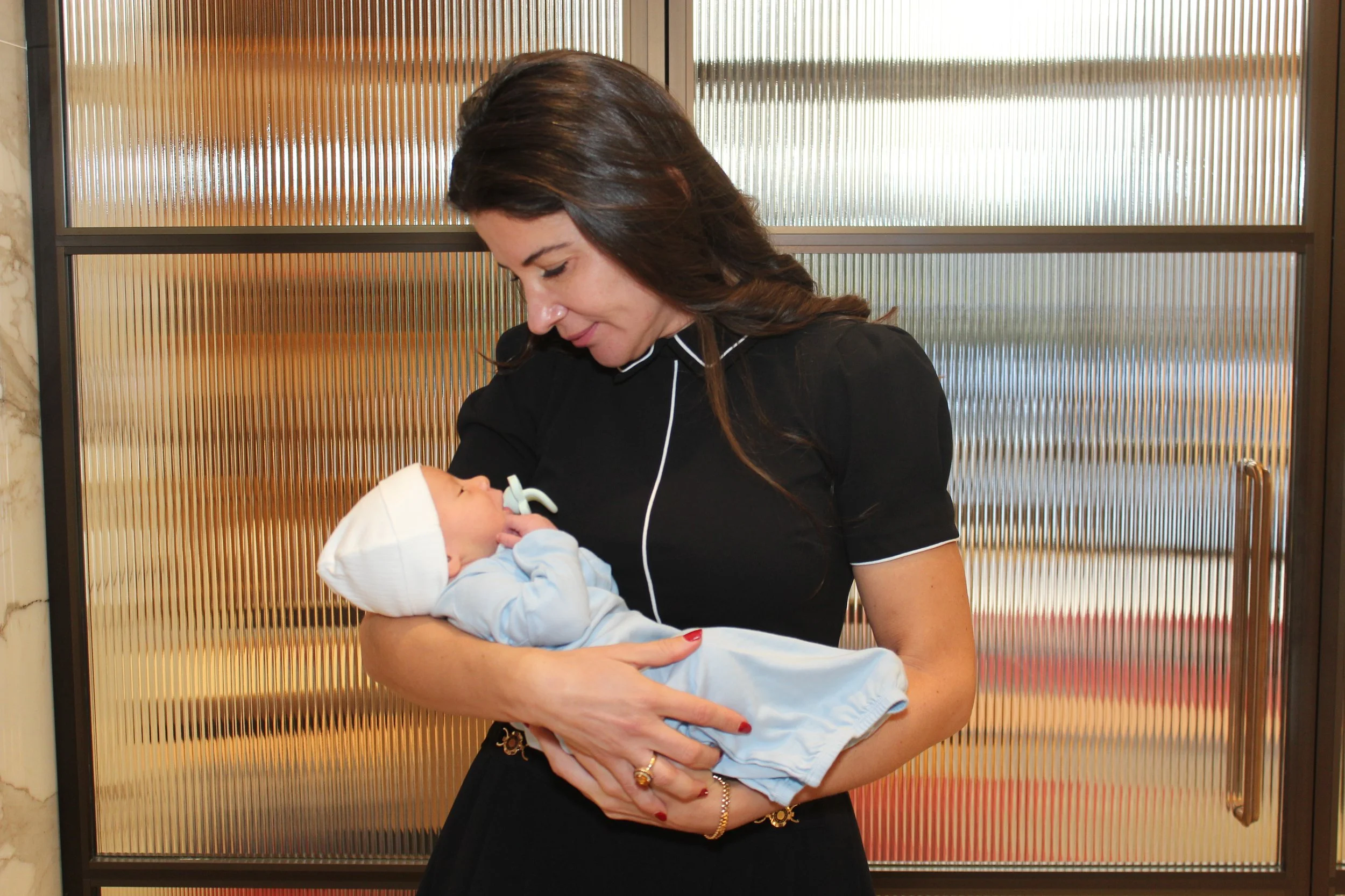 Woman in a black dress gently holding a newborn baby in a light blue outfit and white hat.