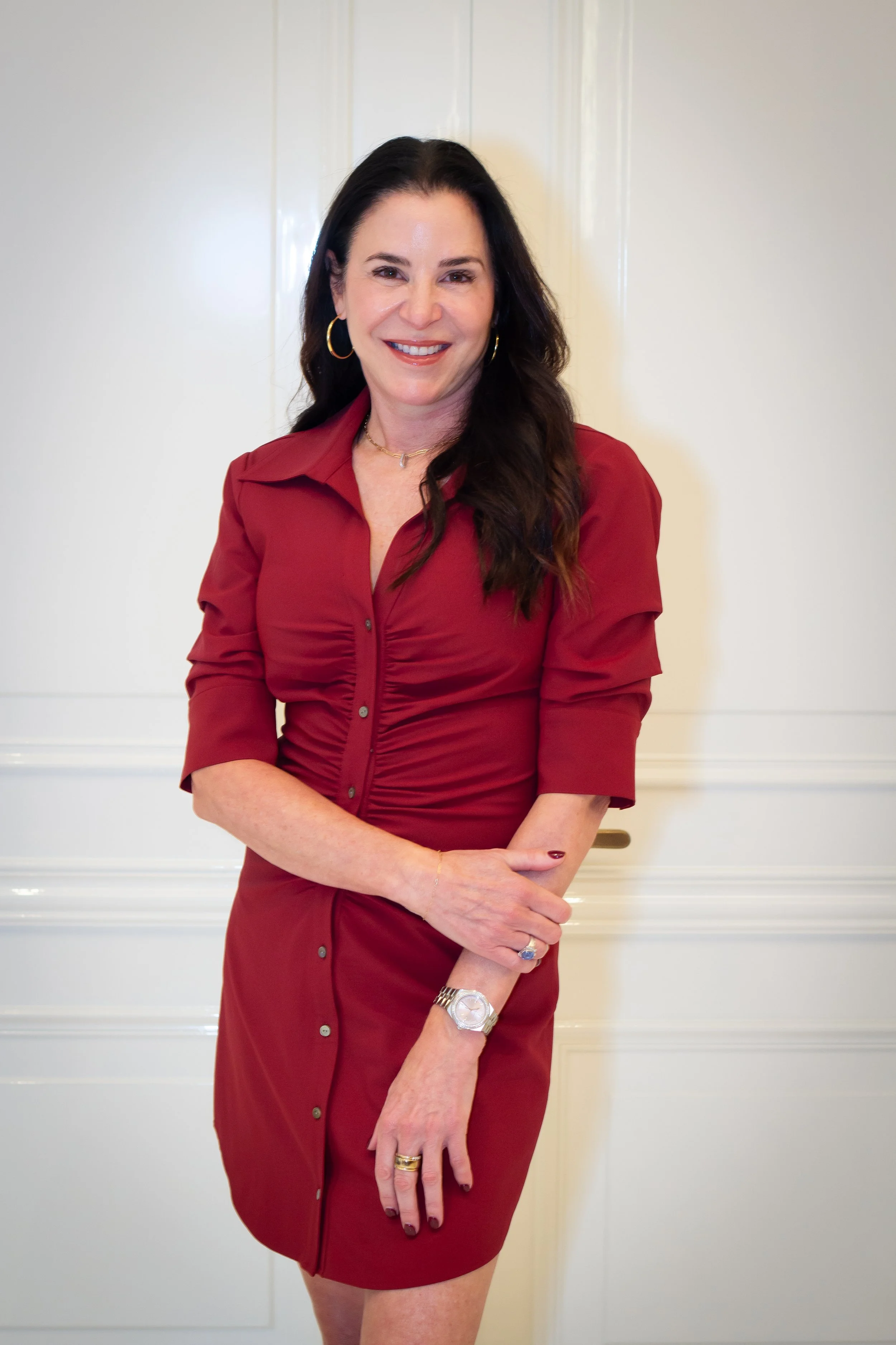 Smiling woman with long dark hair wearing a burgundy dress and gold hoop earrings, standing indoors