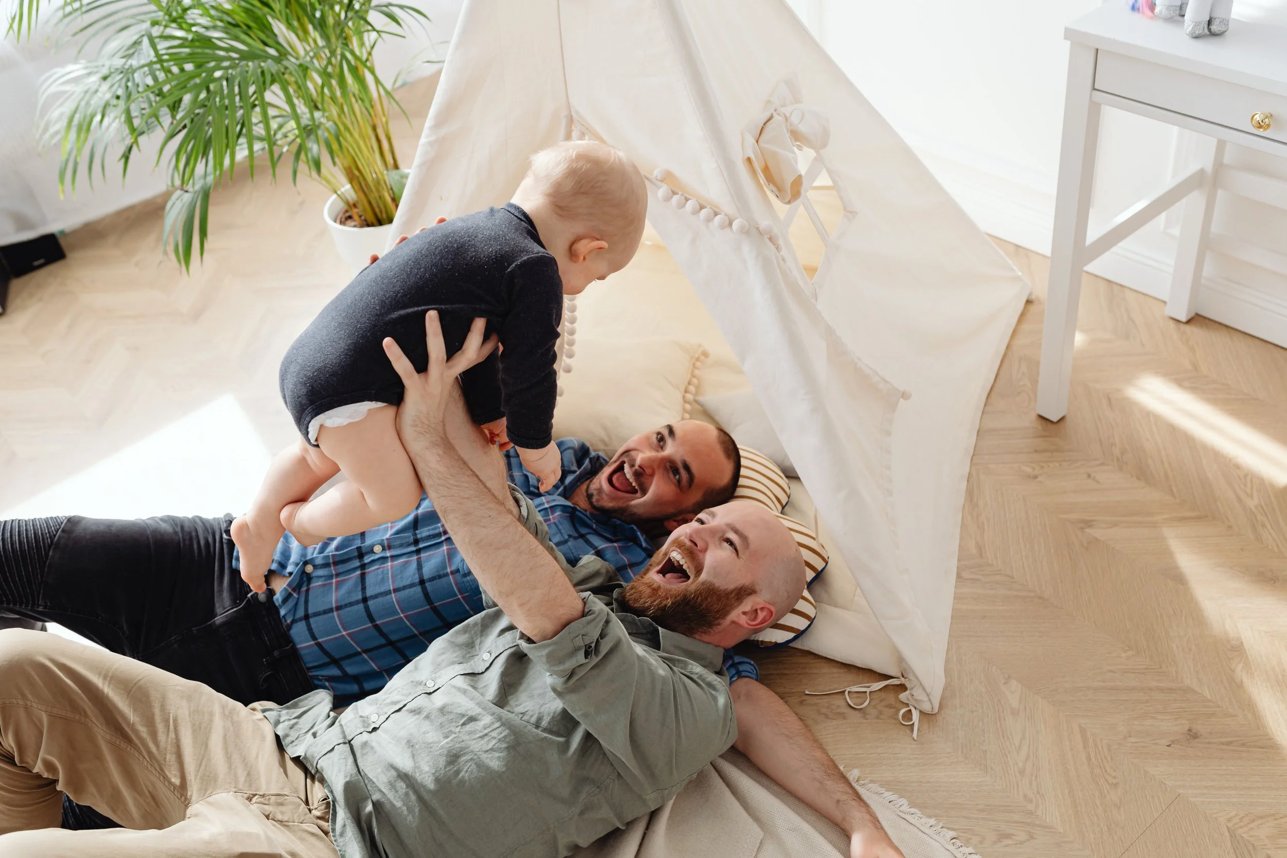 Two smiling fathers play with their baby on the floor beside a cozy indoor teepee tent.