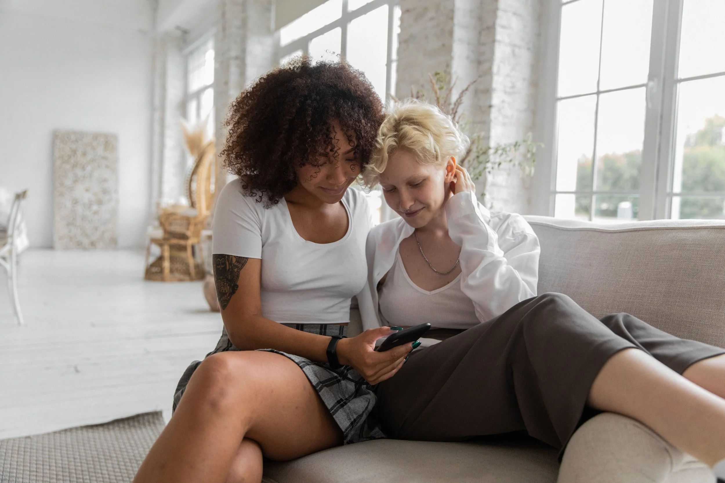 Two women sitting closely on a couch, looking at a smartphone in a bright, sunlit living room