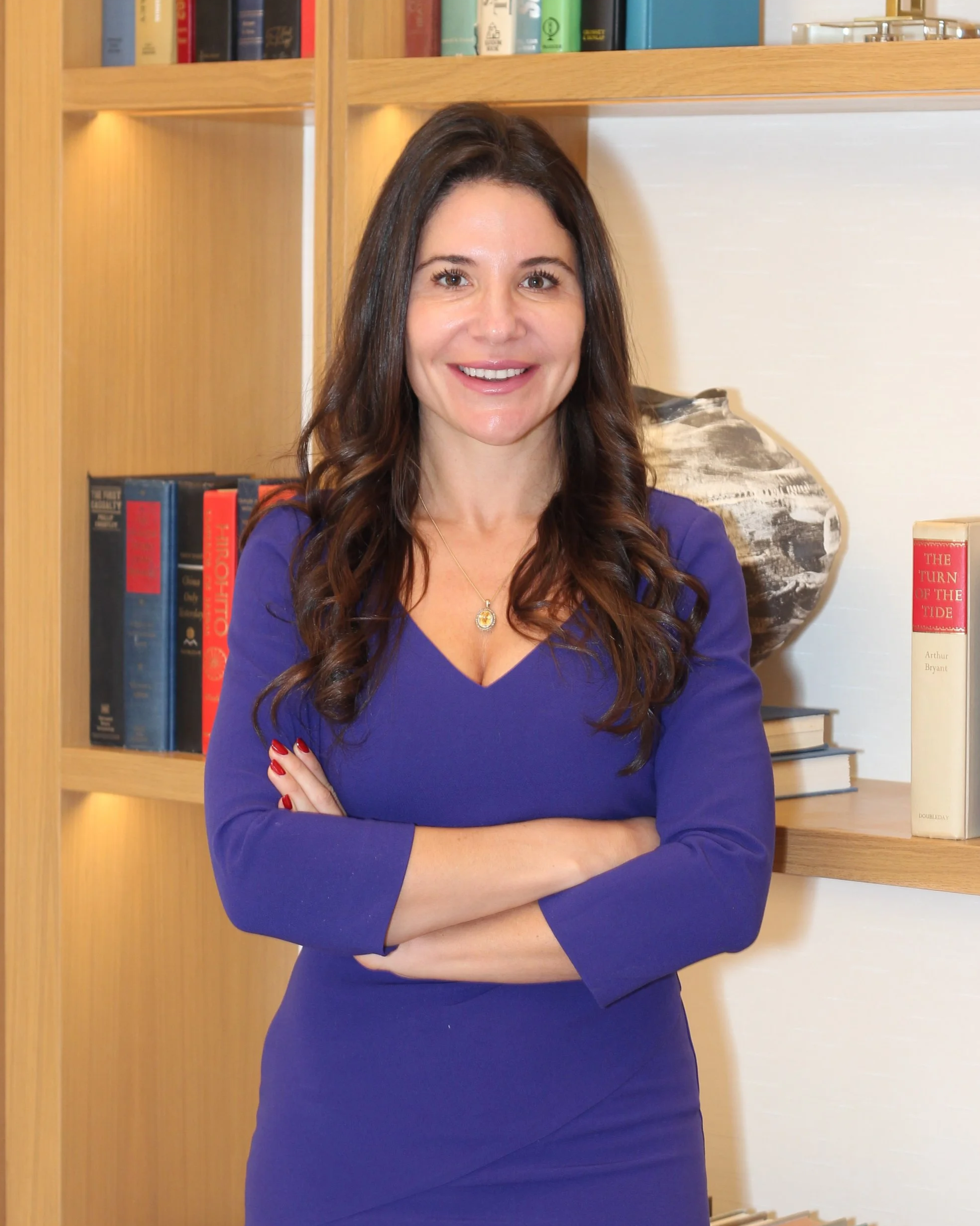 Smiling woman in a purple dress standing with crossed arms in front of a bookshelf