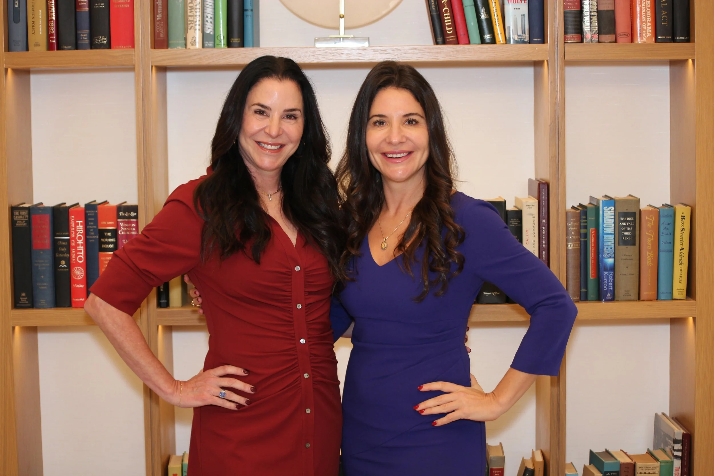 Two smiling women in red and blue dresses pose in front of a bookshelf
