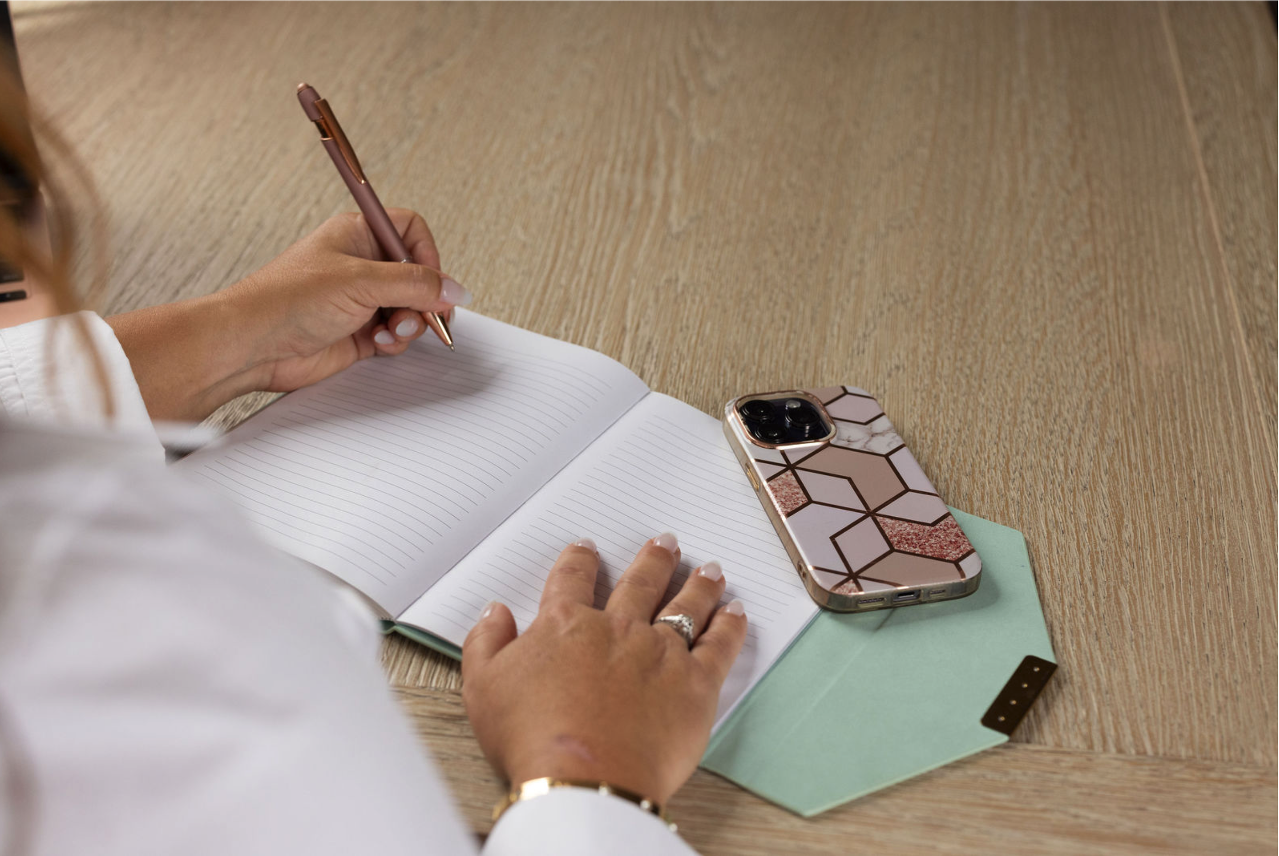 Person writing in an open lined notebook beside a smartphone on a wooden desk