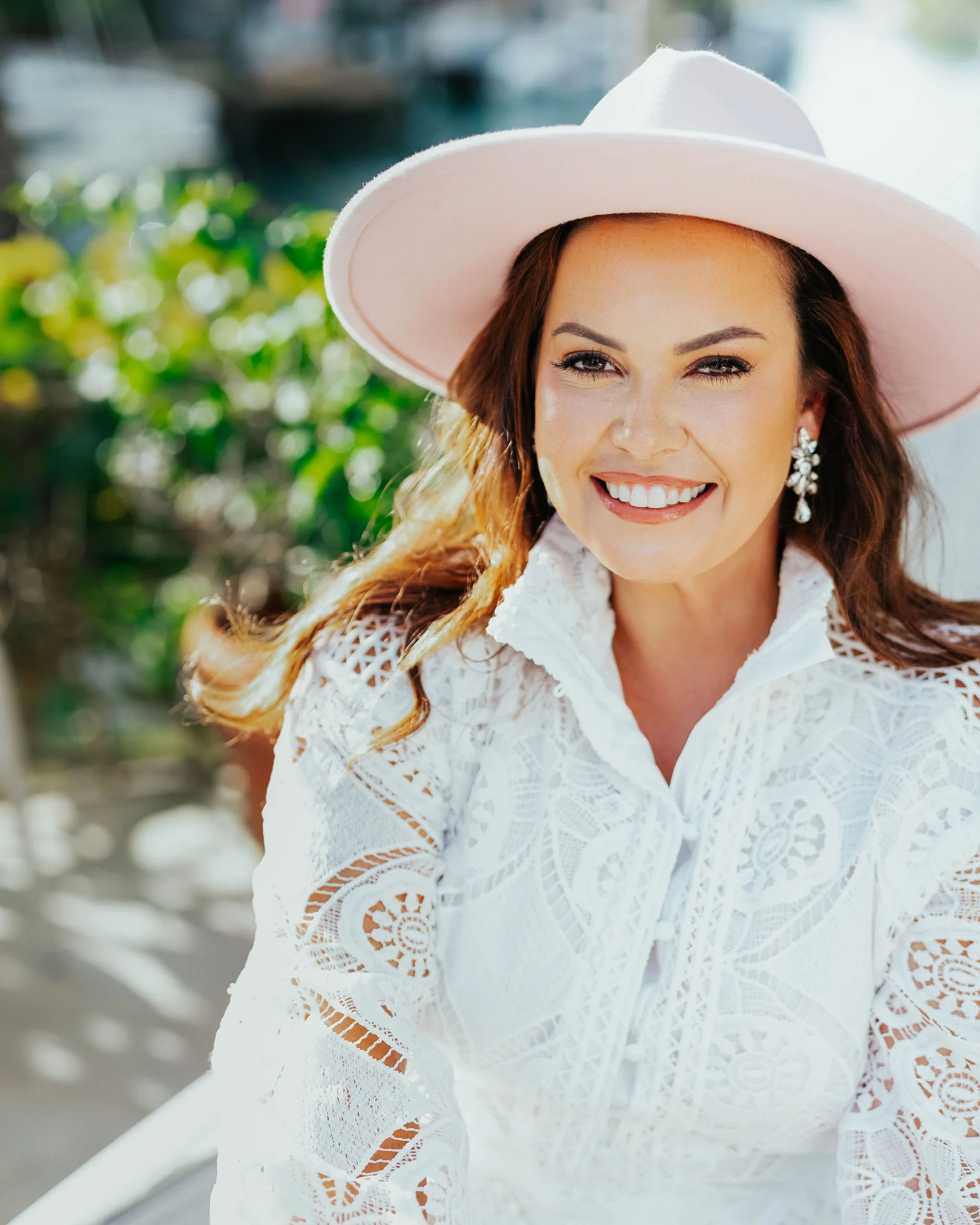 Smiling woman wearing a wide-brimmed pink hat, white lace blouse, and dangling earrings outdoors