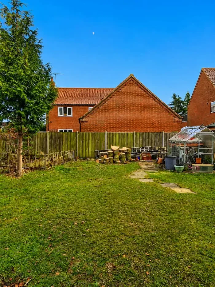 Backyard with grass, a tree, a wooden fence, a greenhouse, and a brick house with a red tiled roof under a clear blue sky.