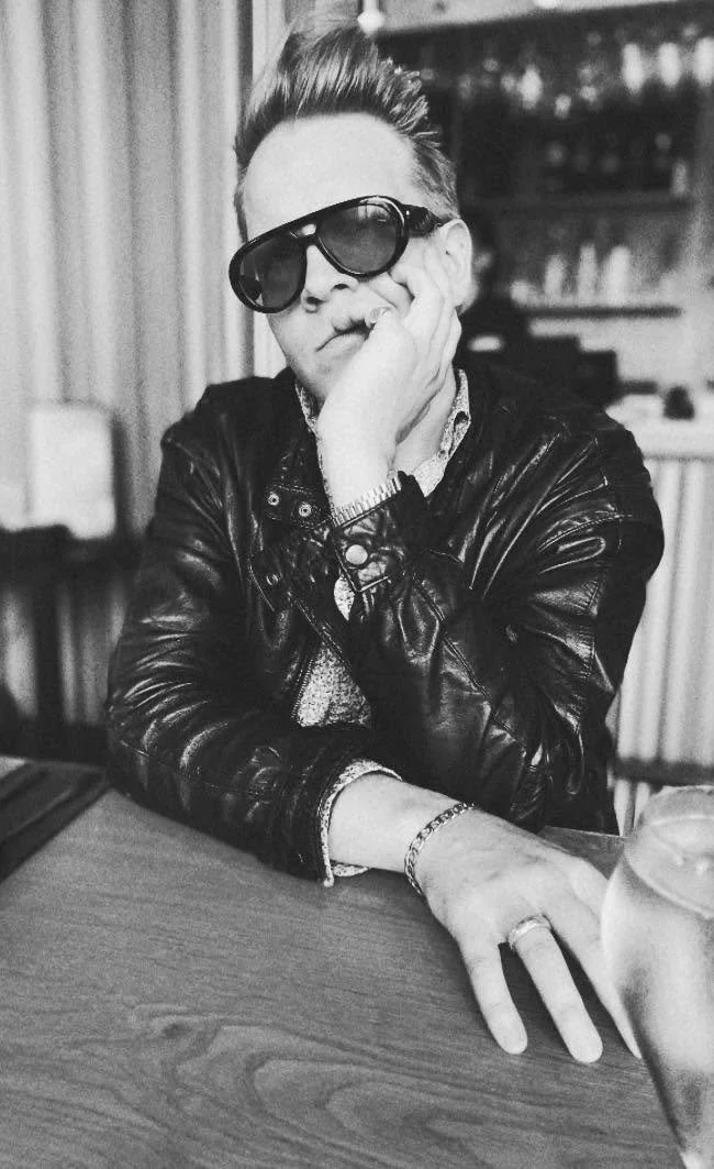 A person with styled hair wearing large sunglasses, a leather jacket, and jewelry, sitting at a table with a drink in front. They are resting their face on their hand in a relaxed pose.