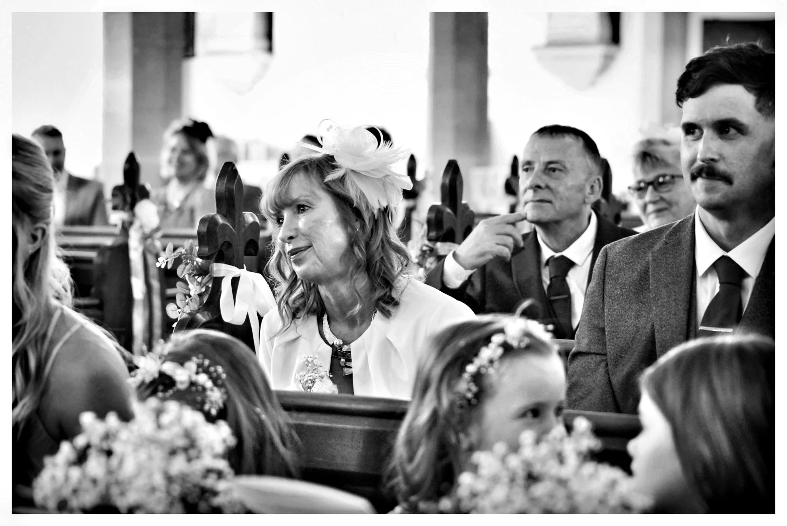 Black-and-white wedding ceremony photo showing guests seated in a church pew, including a woman wearing a large fascinator and a mustached man in a suit.