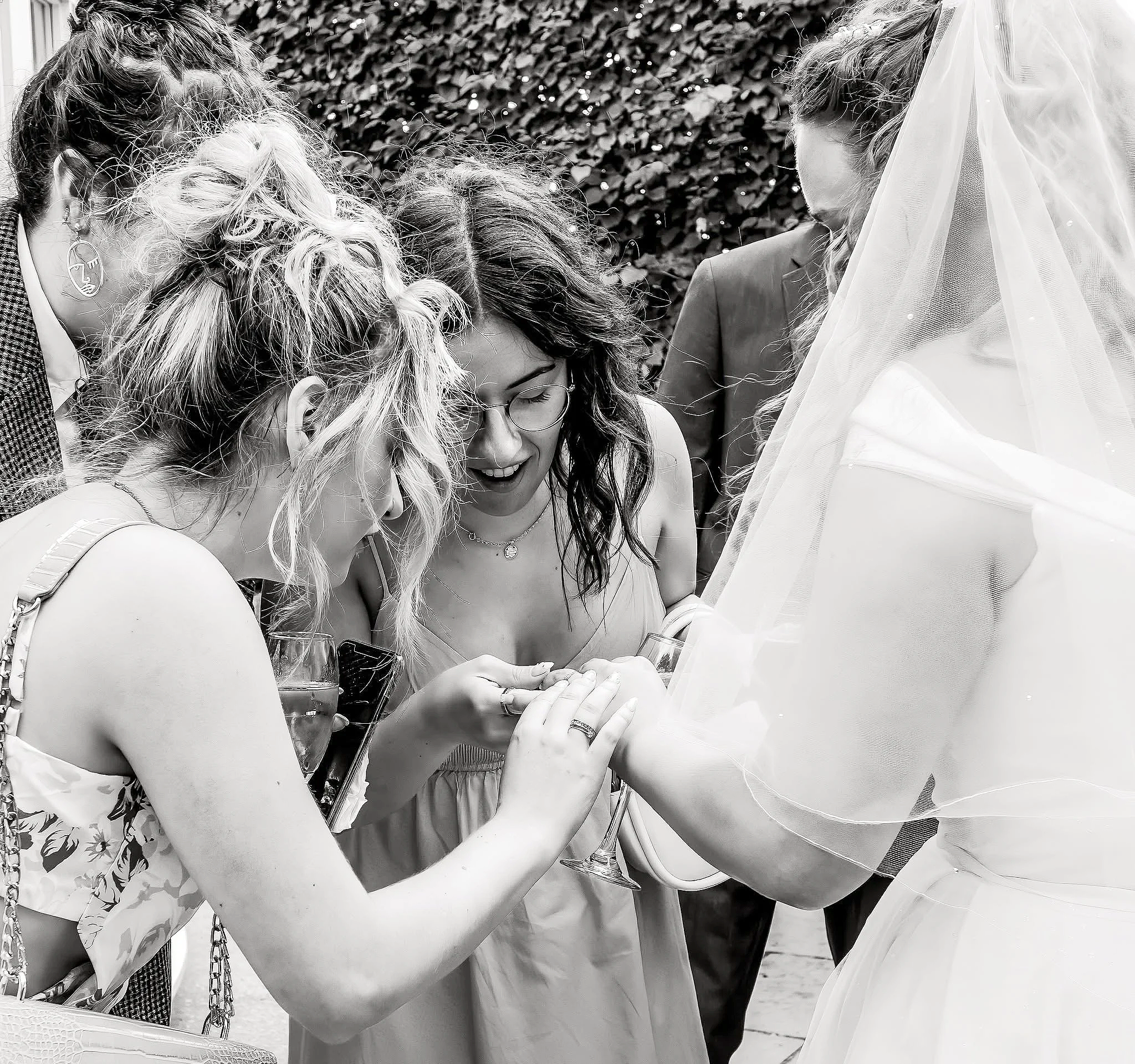 Group of women close together, looking at a smartphone and smiling, outdoors. One woman wearing a veil, suggesting a wedding celebration.
