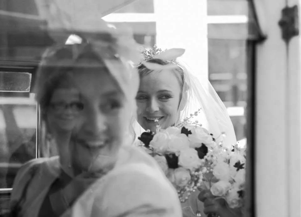 Black-and-white wedding photo of a smiling bride holding a bouquet behind an older woman