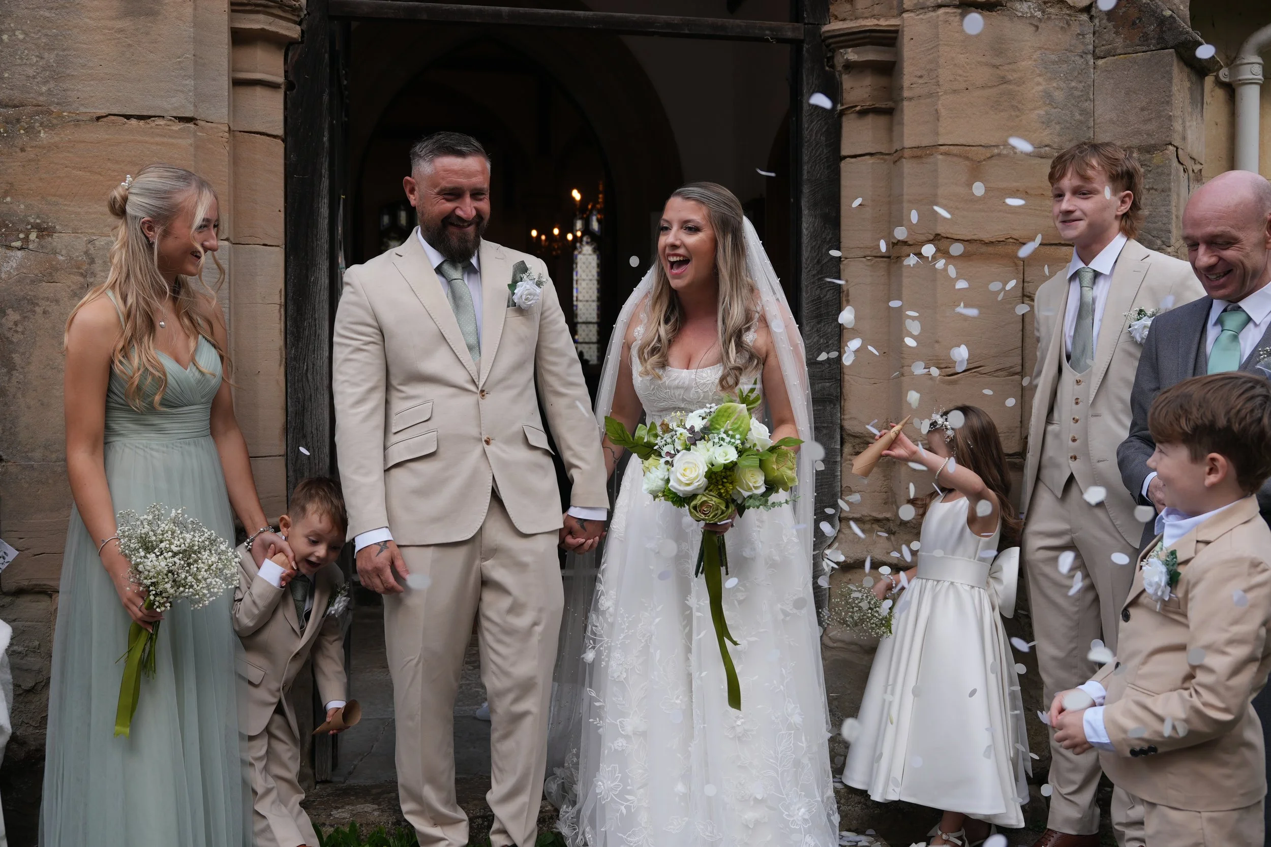Bride and groom leaving a stone church while wedding guests and children toss white confetti.