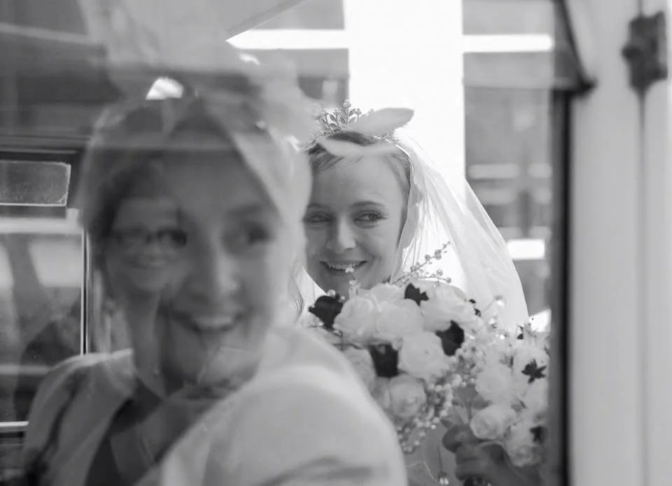 A bride with a veil and floral headpiece holding a bouquet, smiling behind a glass reflection. An older woman, likely a family member, is in the foreground.