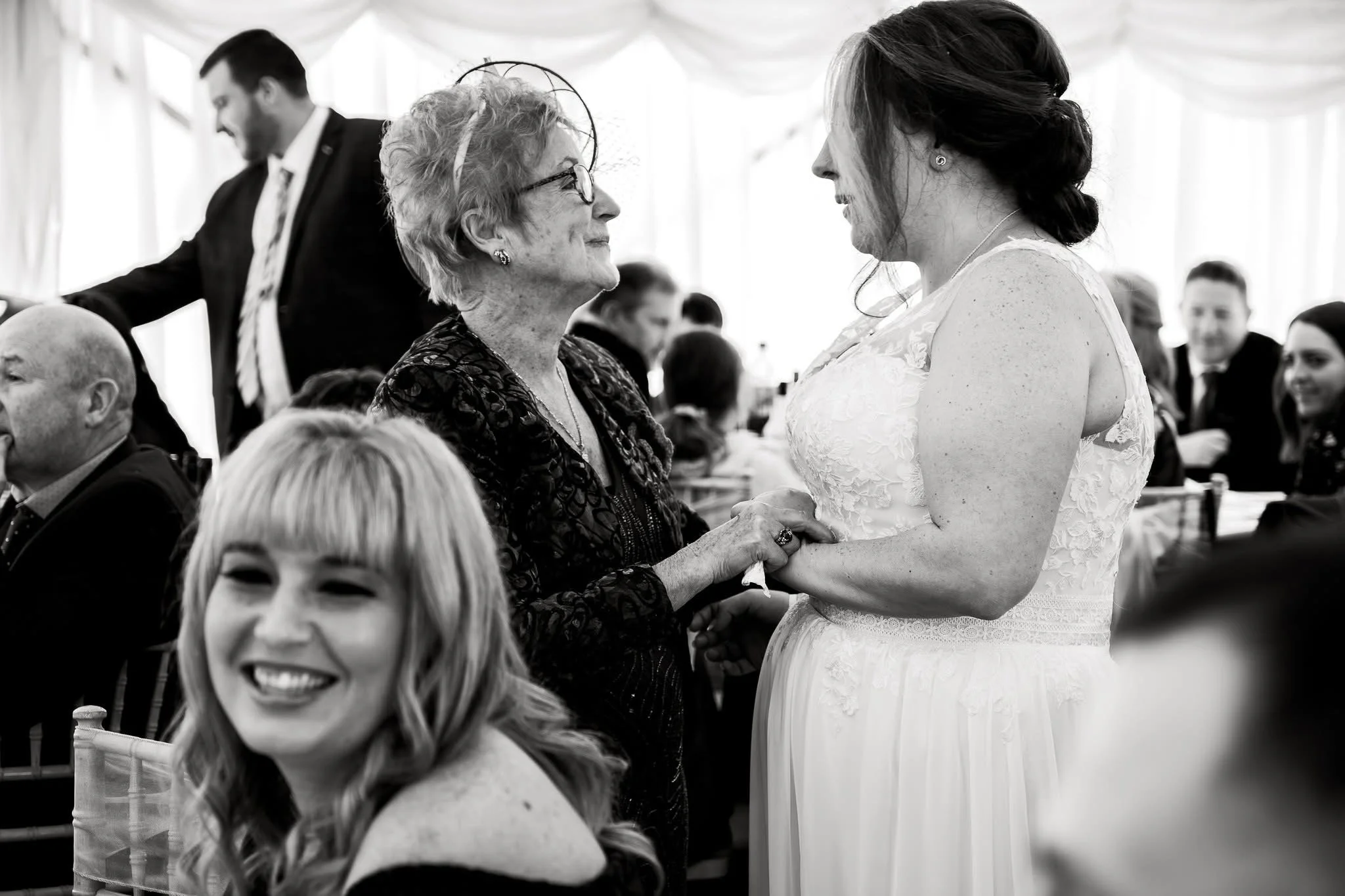 A bride and an elderly woman holding hands and smiling at each other during a wedding reception. The bride is wearing a lace wedding dress, and the elderly woman has glasses and curly hair. The scene takes place in a decorated tent with other guests in the background.