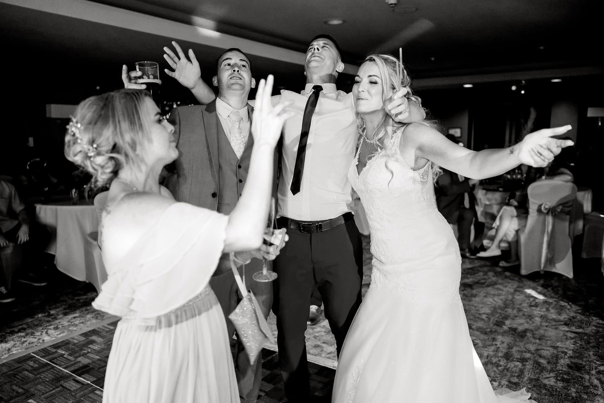 Black and white photo of a wedding celebration with five people dancing and enjoying themselves indoors. The bride in a white gown is in the center, with her eyes closed and a smile, as friends or family surround her with cheerful expressions.