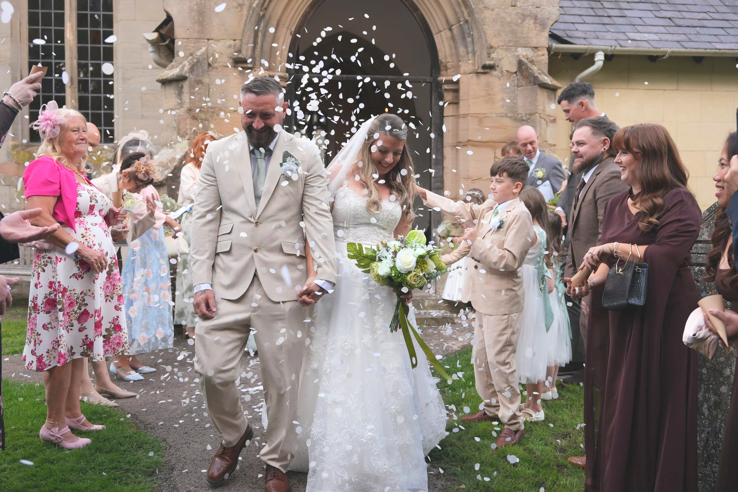 A bride and groom walking hand-in-hand through a shower of confetti outside a church, surrounded by friends and family celebrating their wedding.