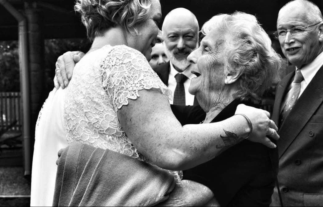 A group of elderly and middle-aged people smiling and embracing each other outdoors at a social gathering.
