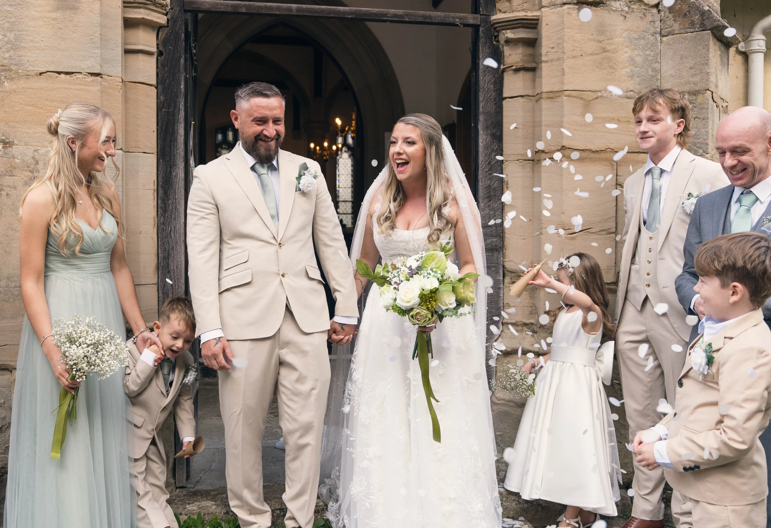 A wedding celebration outside a church with the bride in a white dress holding a bouquet, and the groom in a beige suit, smiling and holding hands. Family and friends surround them, throwing confetti.