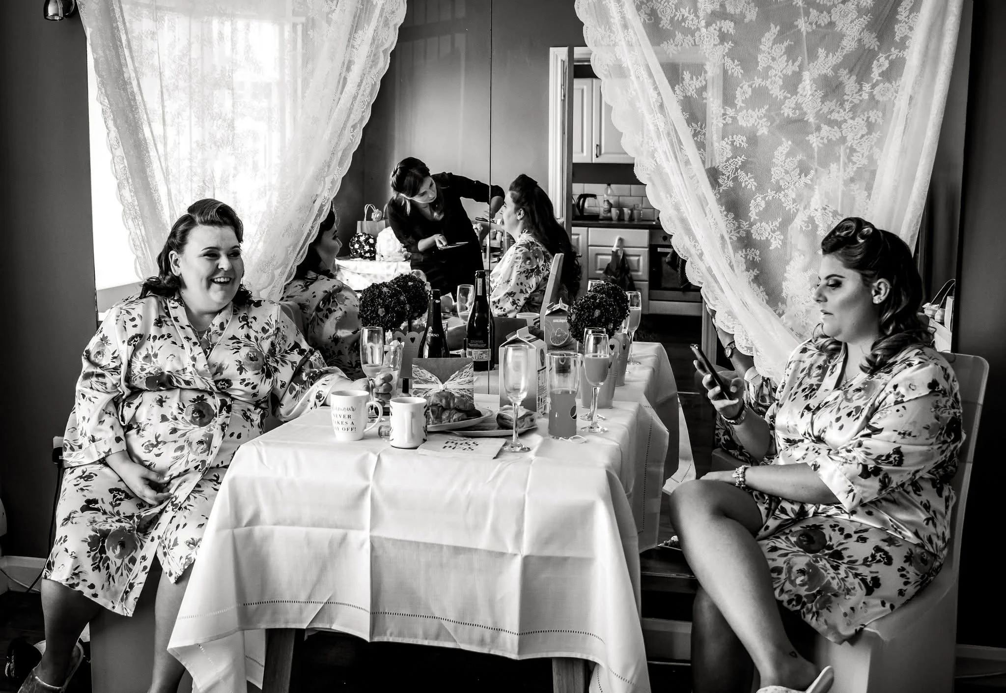 Black-and-white photo of women in floral dresses gathered around a dining table with drinks and food, framed by lace curtains.