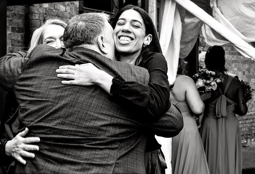 Black-and-white wedding reception photo of three guests embracing and smiling outside a brick building, with bridesmaids in dresses in the background.