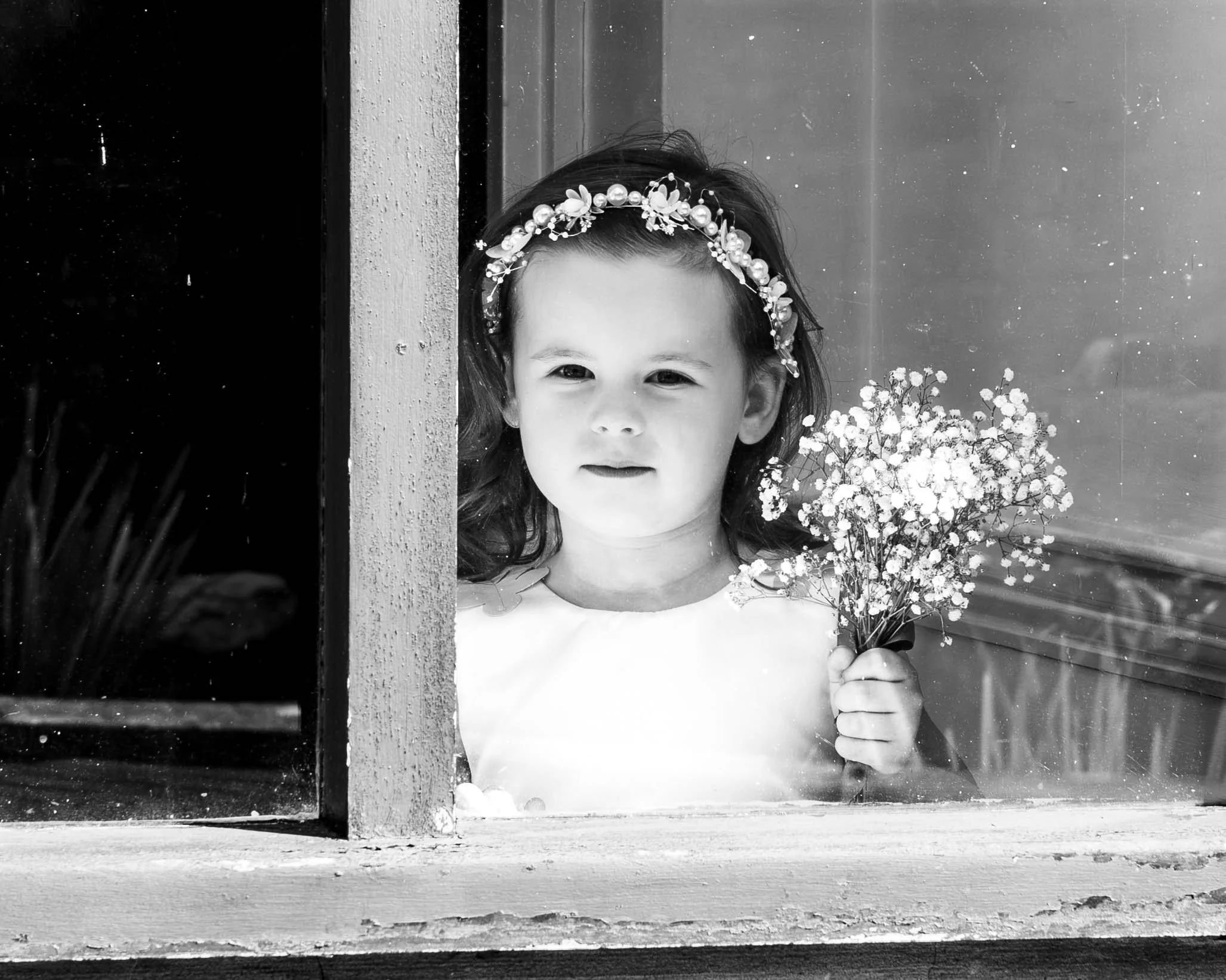 Black-and-white portrait of a young girl wearing a floral headband and holding a bouquet of baby's breath flowers by a window