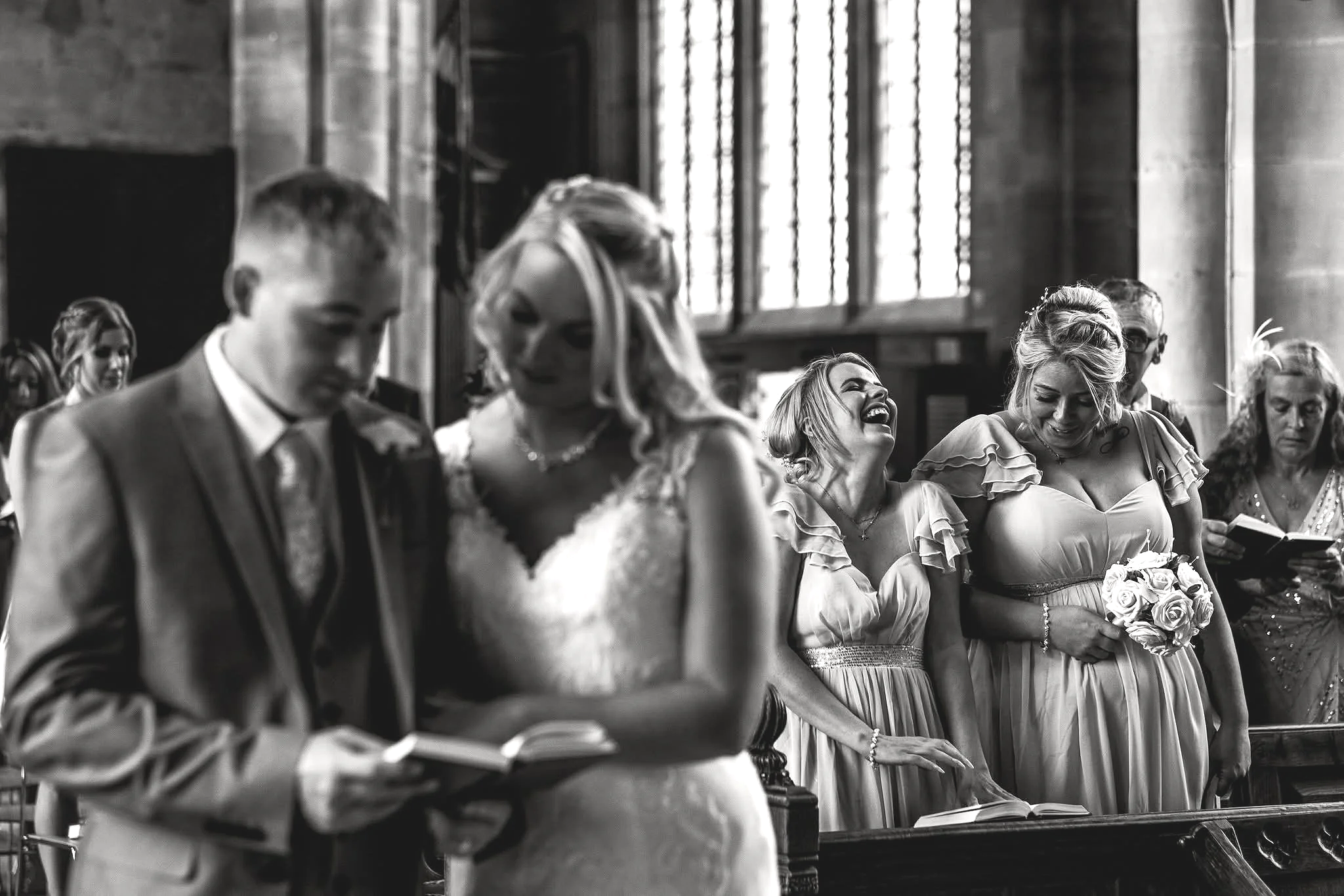 A group of smiling women and men standing together at a wedding ceremony, some holding flowers, inside a church with large windows.