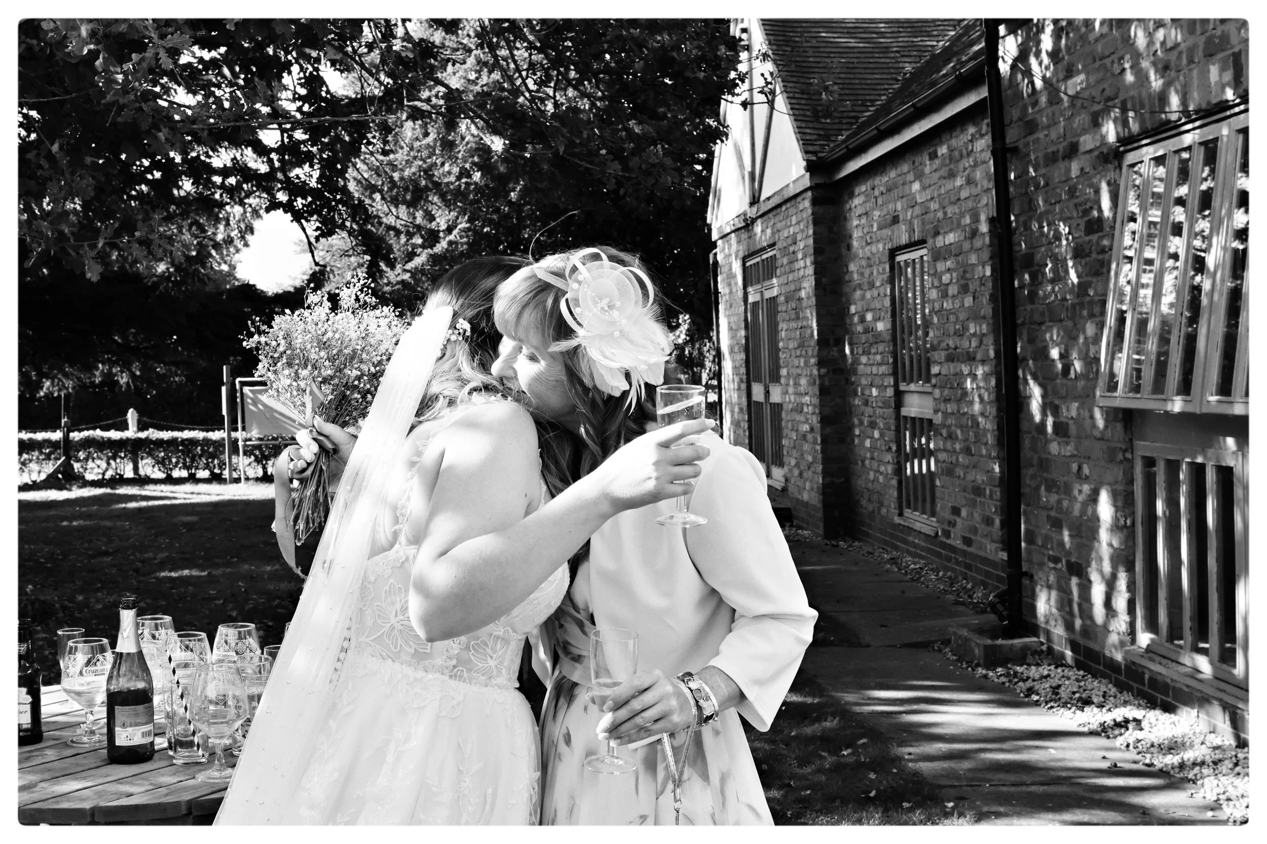 Black-and-white wedding photo of a bride and bridesmaid embracing outdoors beside a brick building, holding flowers and champagne glasses.