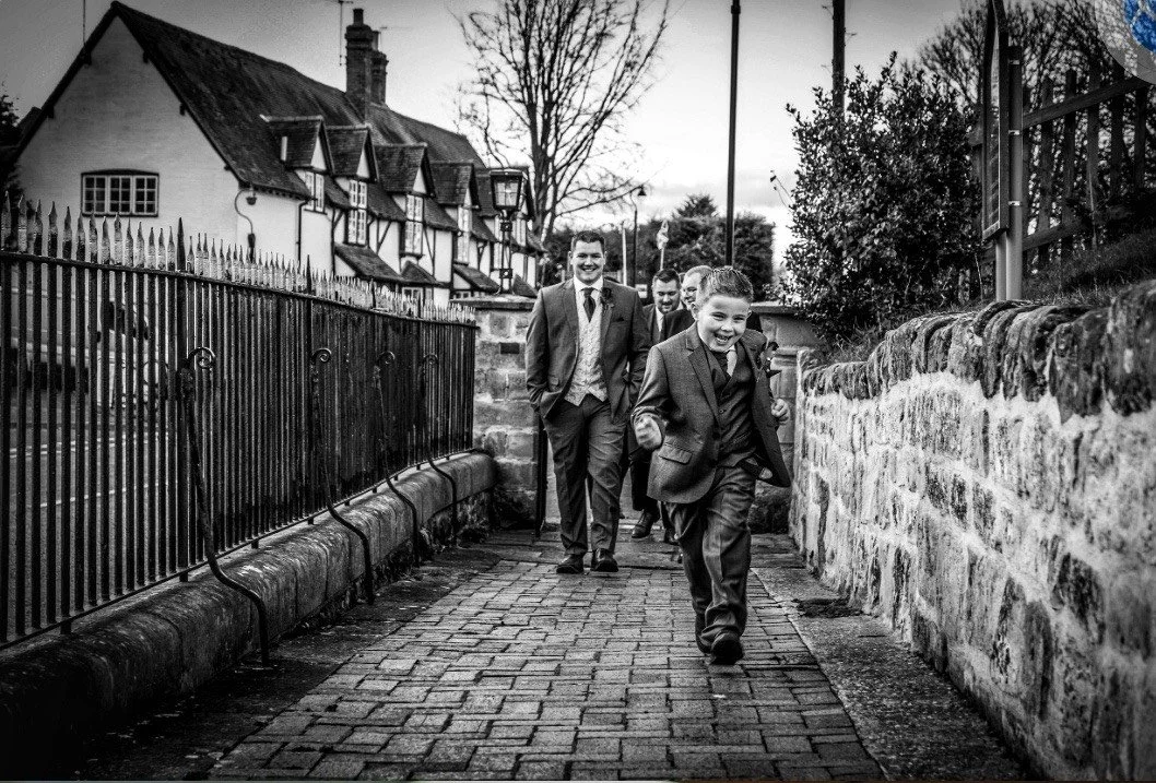 Black-and-white photo of formally dressed boys walking along a cobblestone lane beside historic English cottages and stone walls.