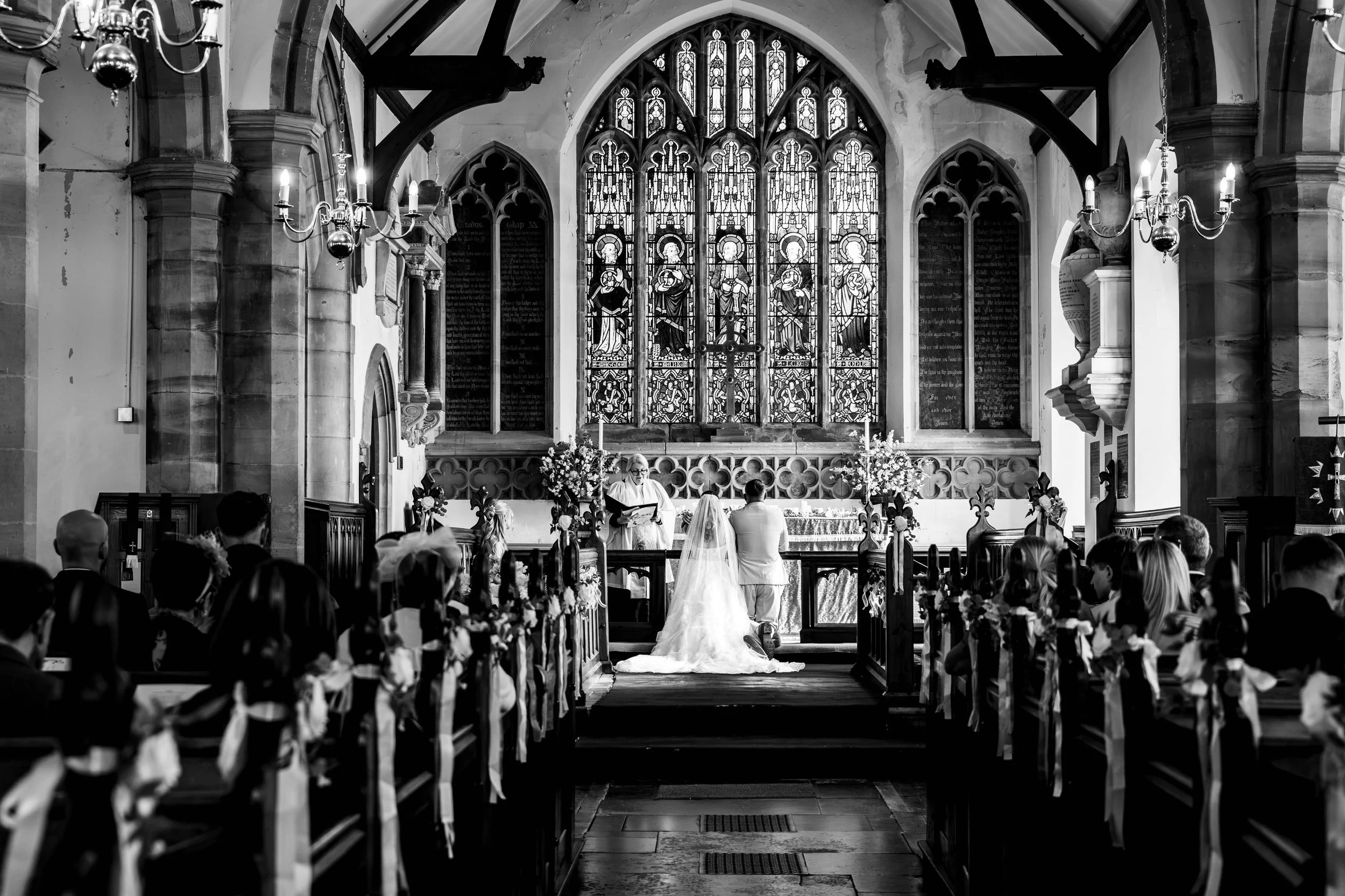 Black-and-white wedding ceremony in a historic church with stained-glass windows, bride and groom at the altar, and guests seated in decorated pews