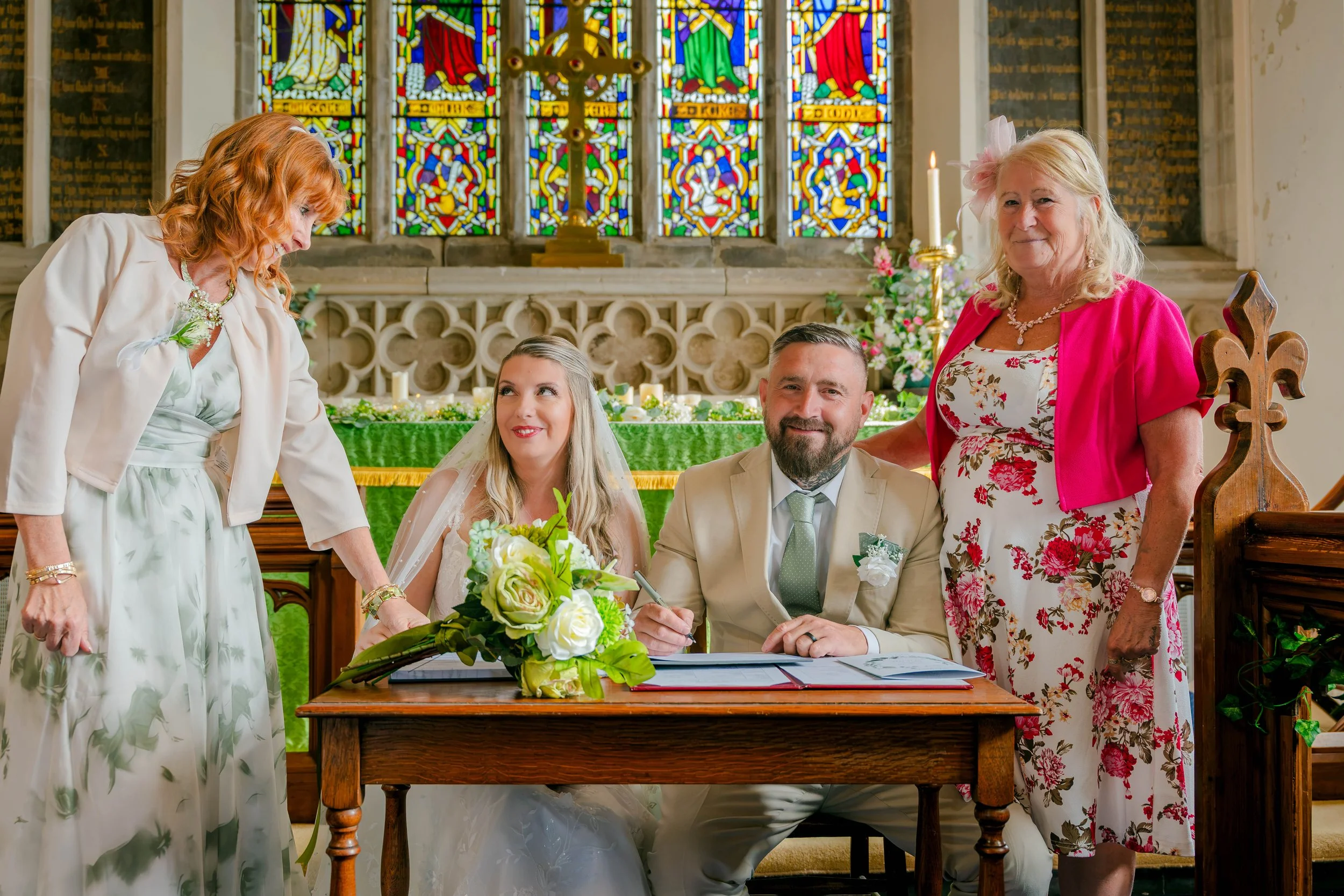 Bride and groom sign wedding papers at a church altar with family members, stained-glass windows, flowers, and candles.