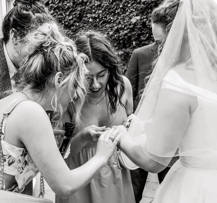 Black-and-white wedding photo of friends helping a veiled bride with her ring outdoors