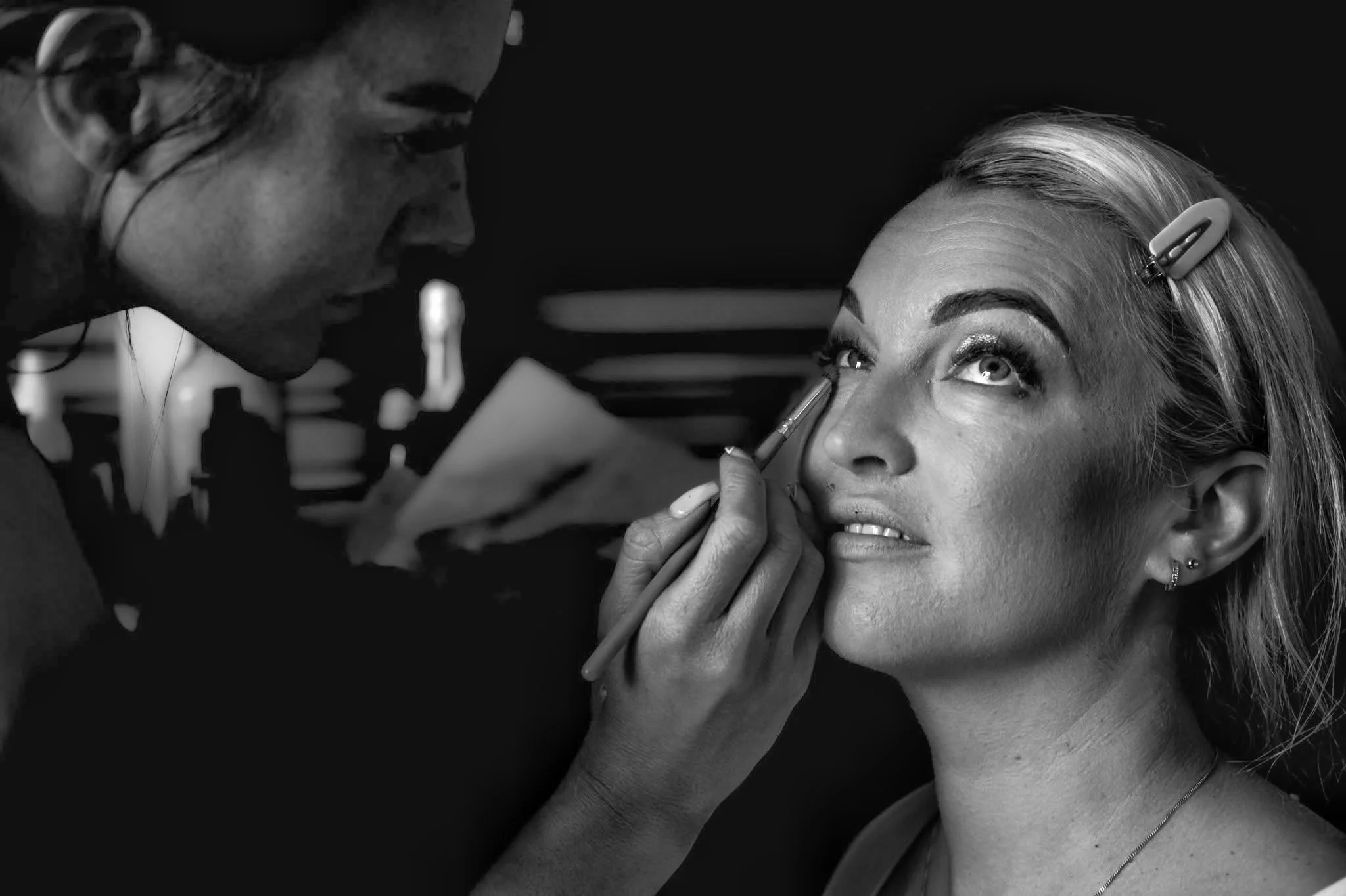 Makeup artist applying eye makeup on a woman with light hair, styled with a hair clip, under professional lighting.