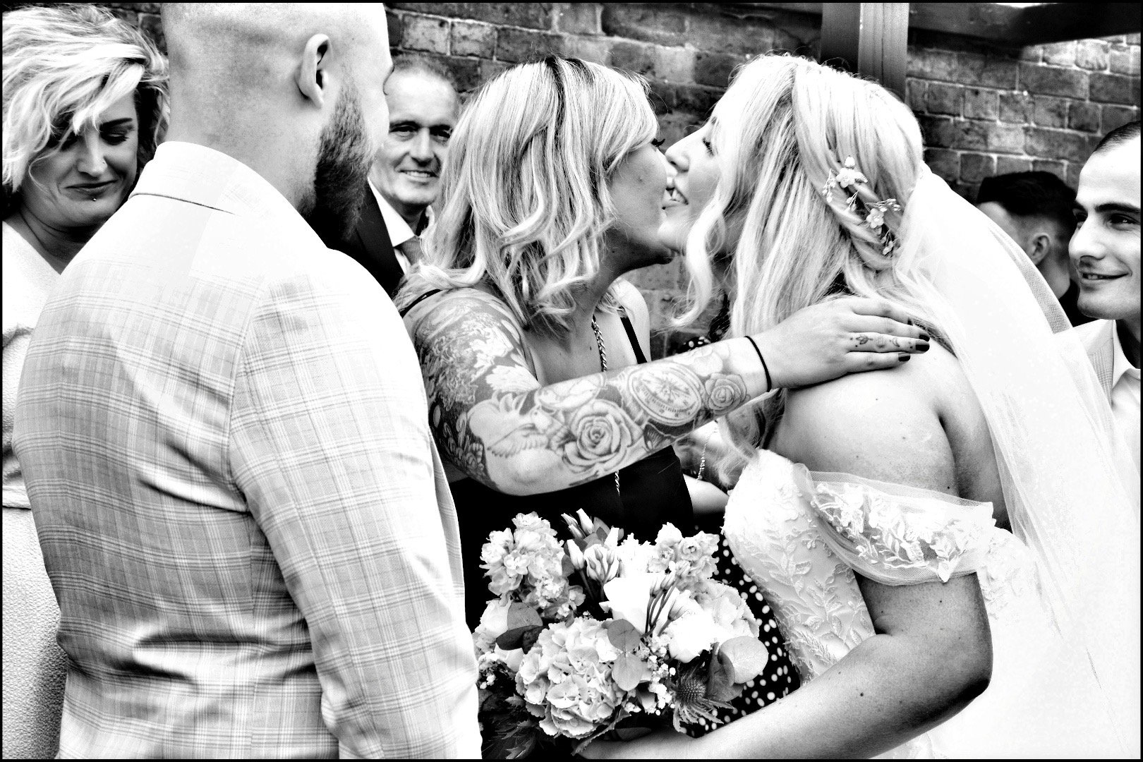 Black-and-white wedding photo of a bride kissing a tattooed bridesmaid while guests gather around them.