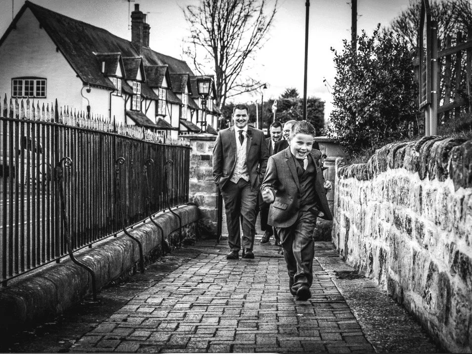 Black-and-white photo of a smiling boy in a suit walking along a cobblestone path with three men behind him, beside historic timber-framed houses and stone walls.
