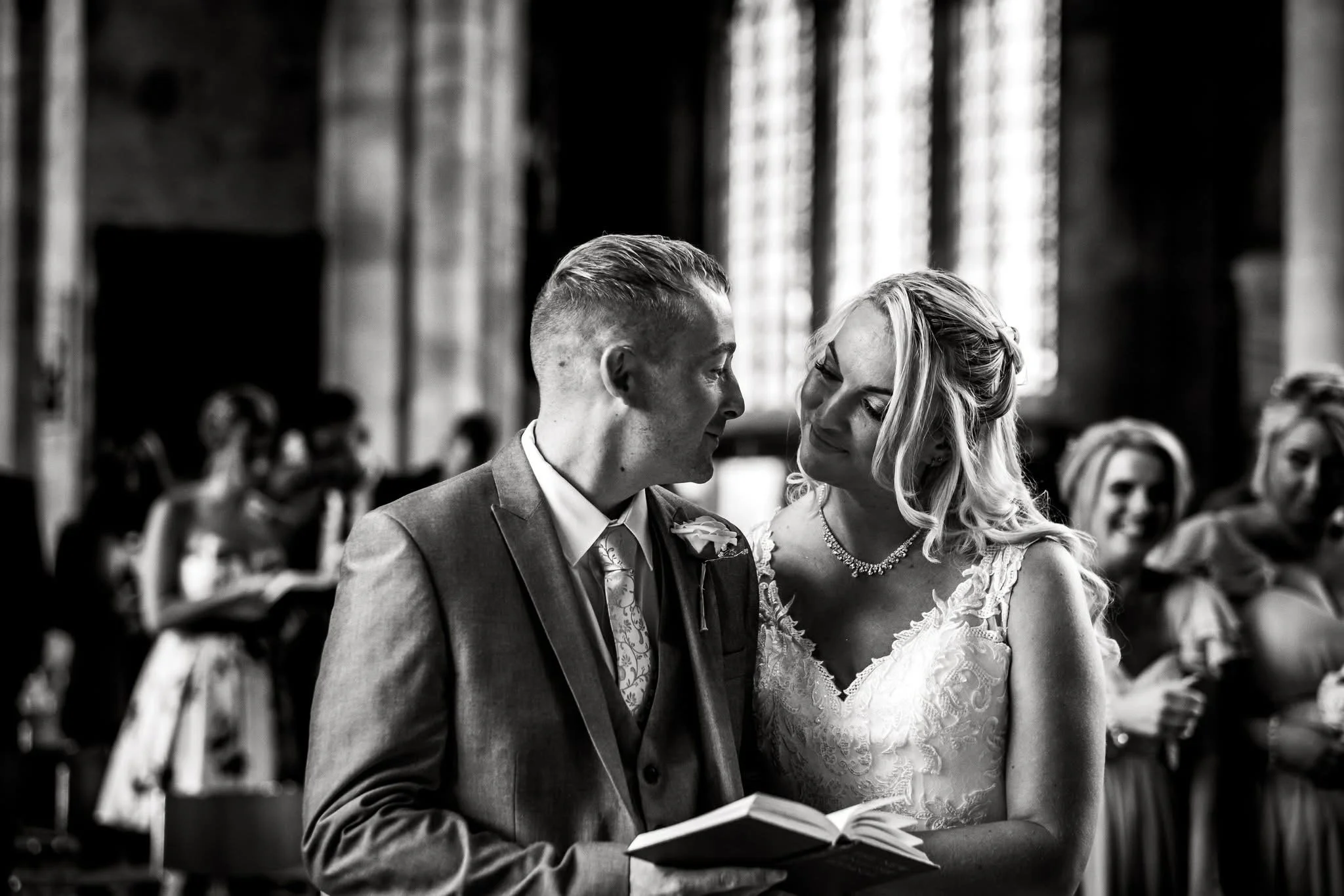 Black and white photo of a wedding ceremony with a bride and groom exchanging vows inside a church. The groom is holding a book, and the couple is looking at each other affectionately.