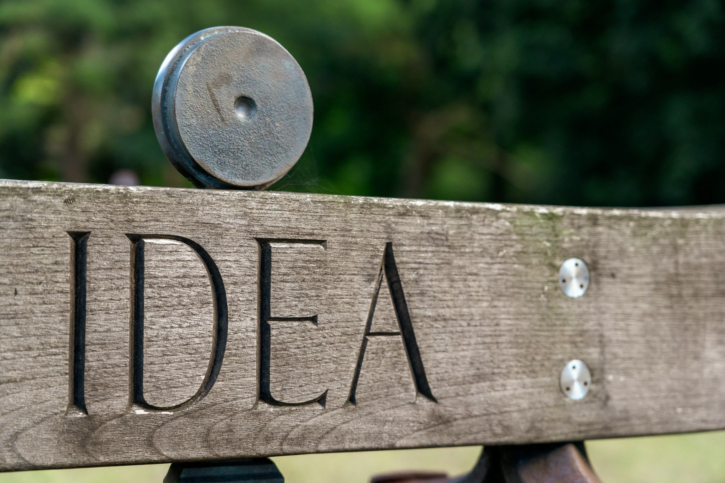 Close-up of a wooden sign with the word 'LOVE' engraved, mounted on a fence, with a round metallic post on top, outdoors with green foliage blurred in the background.