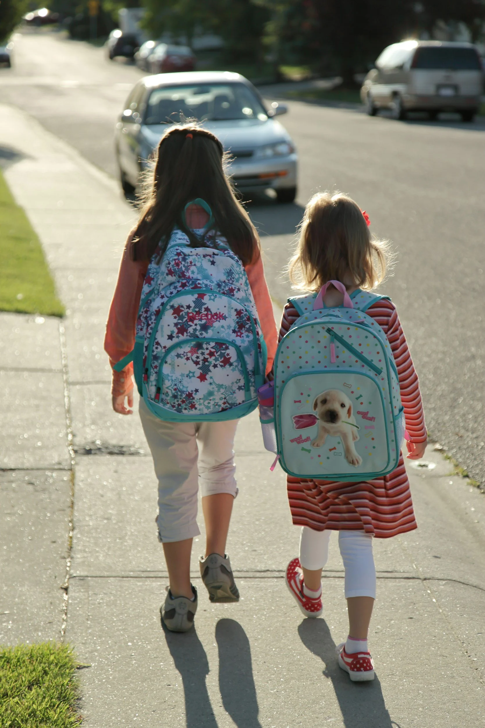 Two young girls walking on a sidewalk with backpacks, one with a colorful star-themed backpack and the other with a backpack featuring a puppy design. They are holding hands, and the sunlight casts their shadows on the pavement.