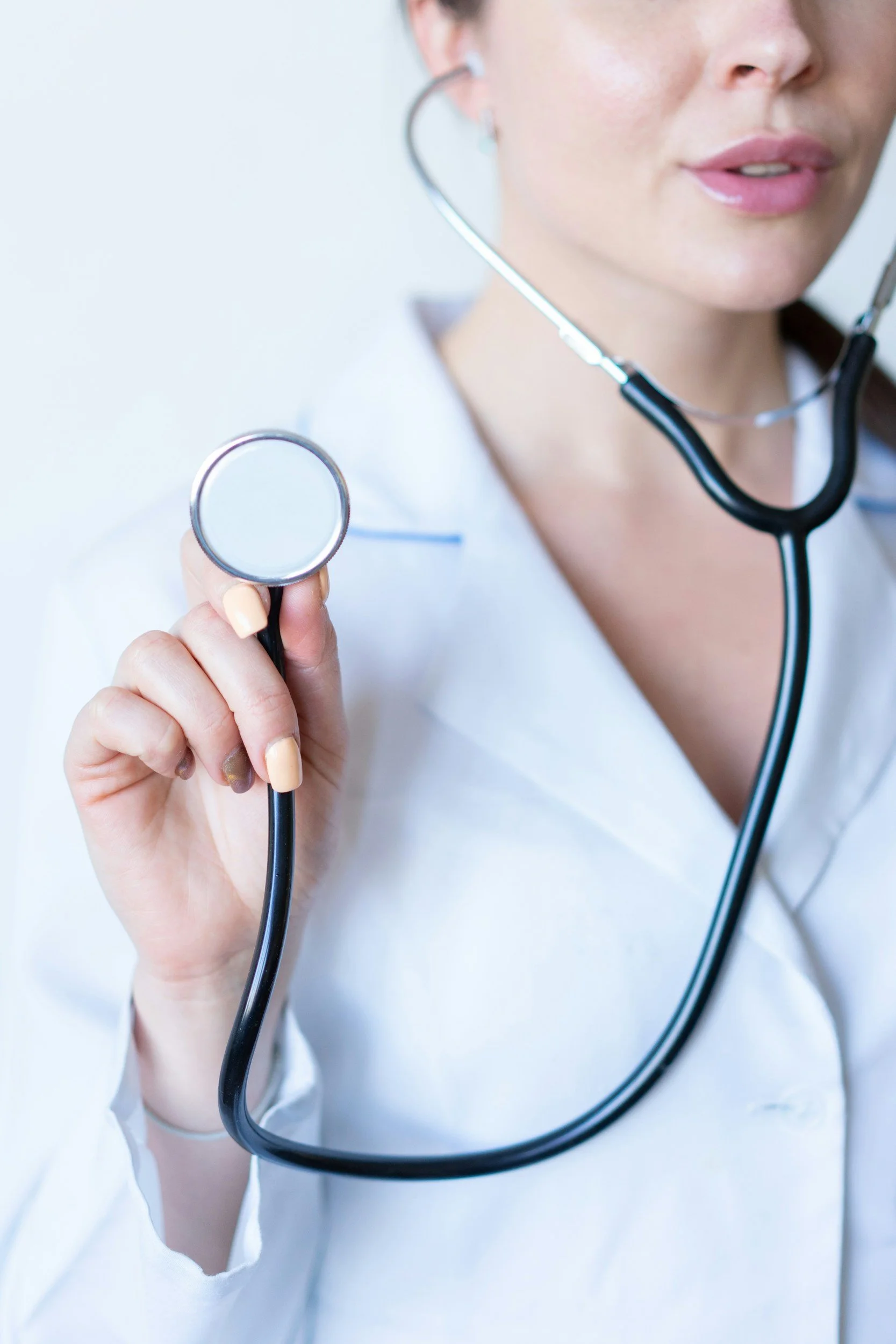 Close-up of a healthcare professional holding a stethoscope in their hand, wearing a white coat, with focus on the professional's face and part of their body.