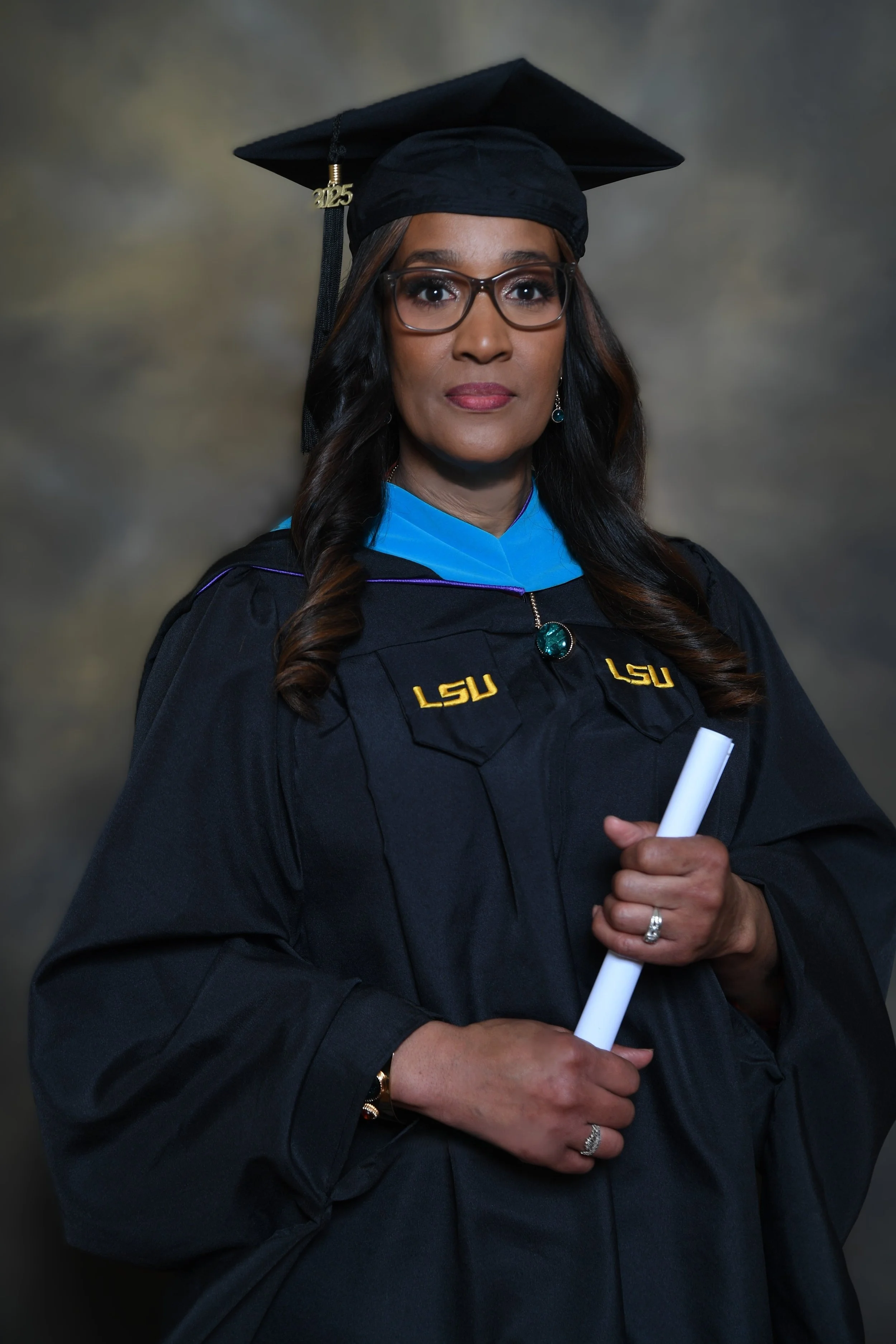 A woman wearing graduation cap and gown with LSU logo, glasses, and jewelry, holding a diploma, against a gray background.