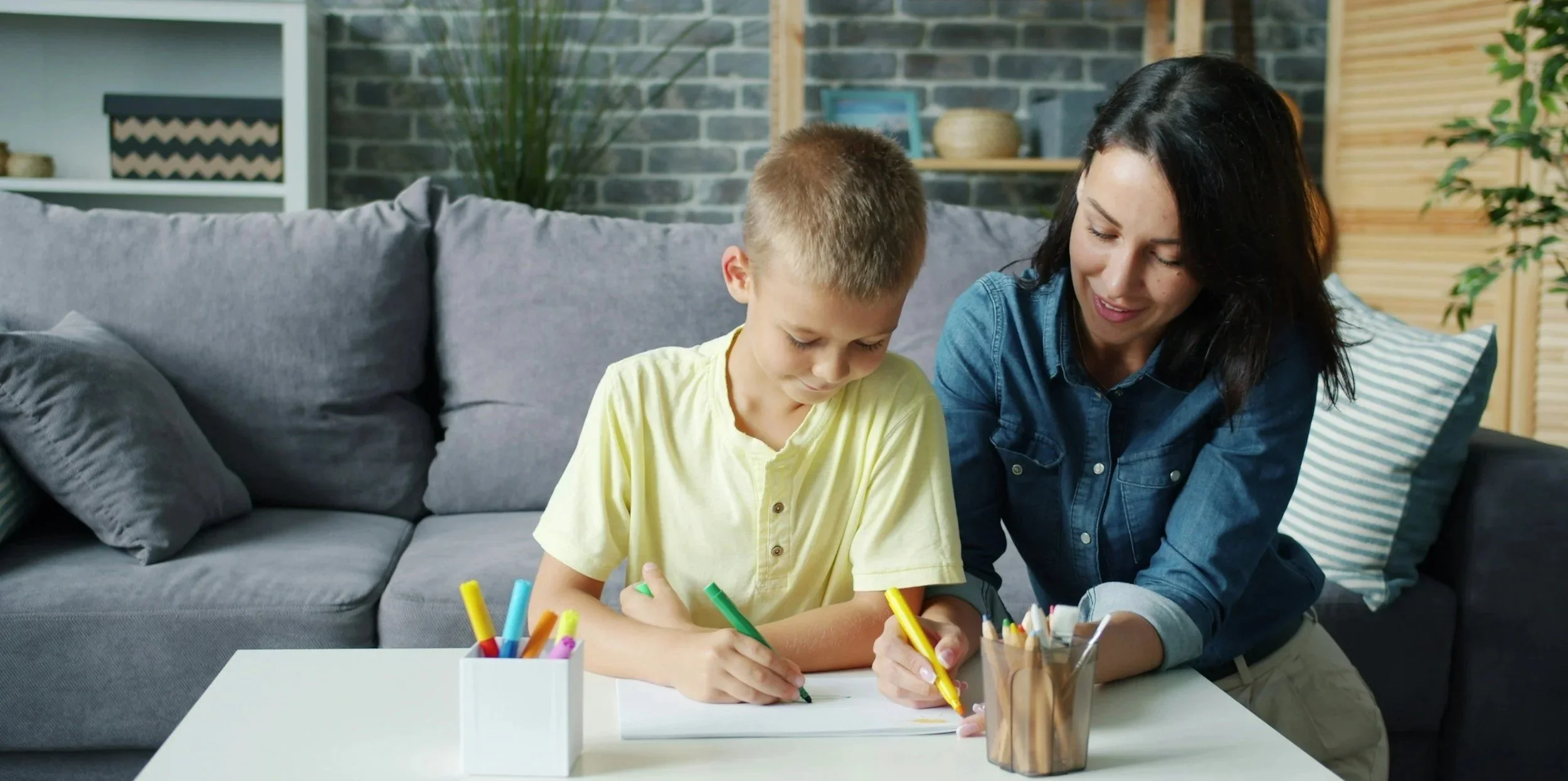 A woman and a young boy sitting on a gray couch, drawing with colorful markers at a white table with a container of markers, in a room with a brick wall and wooden shelves.
