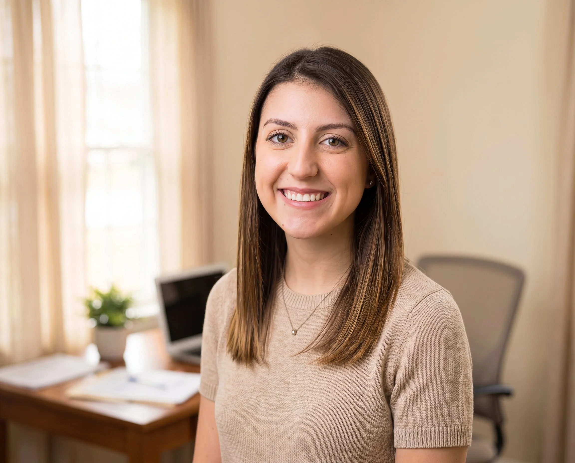 A young woman with brown hair and a beige sweater smiling in an office setting with a desk, computer, and a plant in the background.