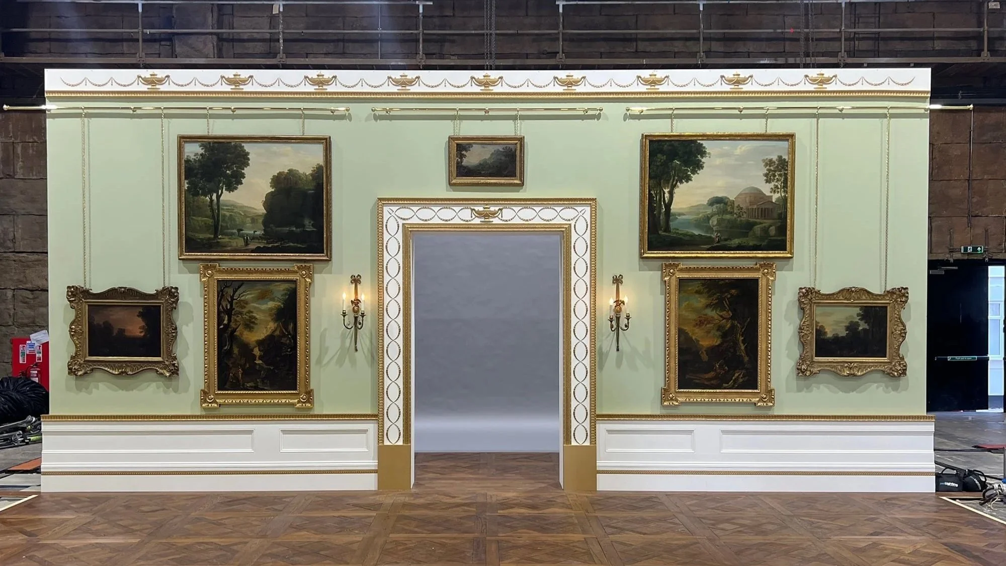 An indoor exhibition wall displaying multiple landscape paintings in ornate gold frames with decorative wall sconces, set against a pale green wall with white and gold trim.