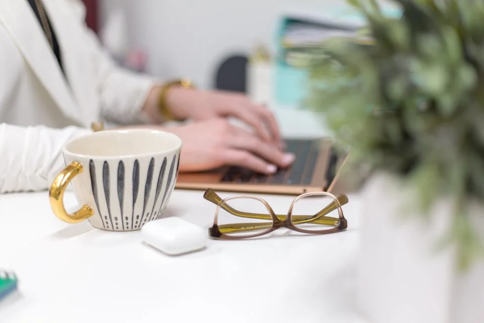 Eyeglasses and patterned coffee cup on a white desk beside a person using a laptop