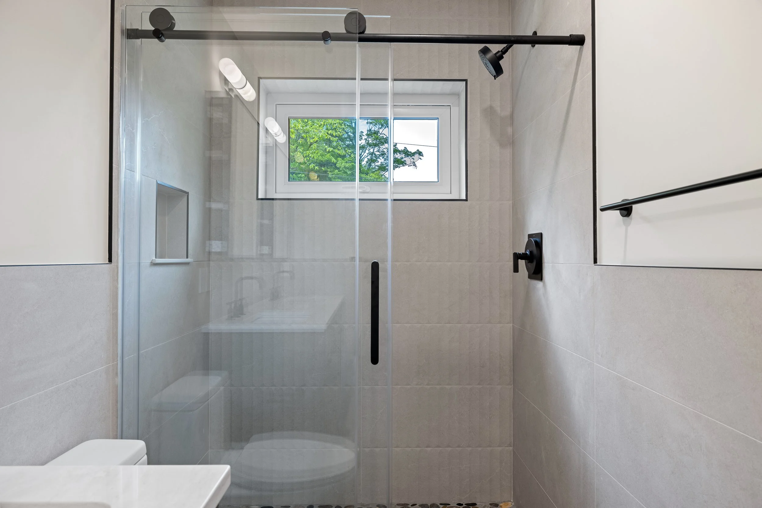 Modern tiled bathroom with a glass-enclosed walk-in shower, black fixtures, recessed shelves, and a window overlooking trees