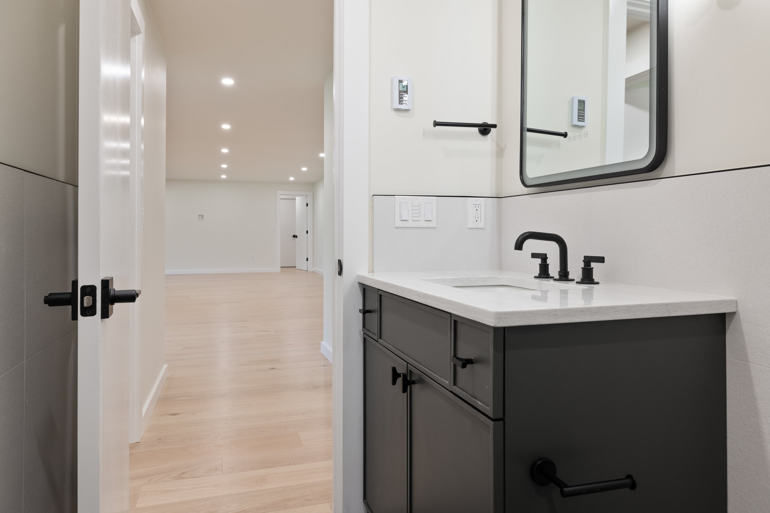 Modern bathroom with matte black vanity, white countertop, black faucet, wall mirror, and open door to a bright hallway