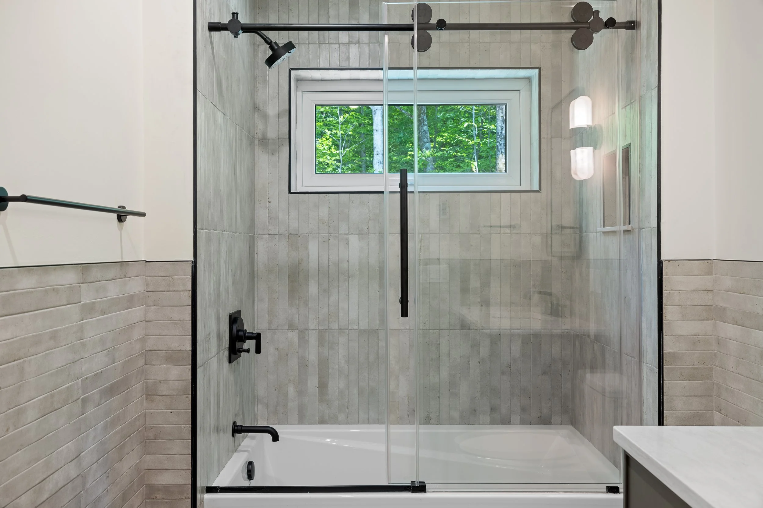 Modern bathroom with a glass-enclosed bathtub shower, black fixtures, gray tile walls, and a window overlooking trees.